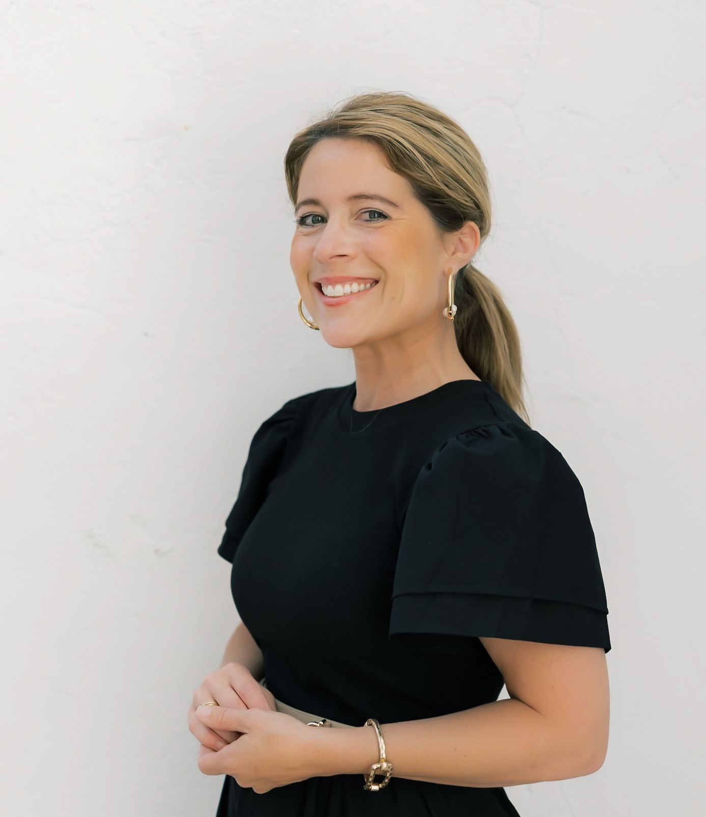 Smiling blonde woman in black dress with ponytail and gold earrings poses against white wall.