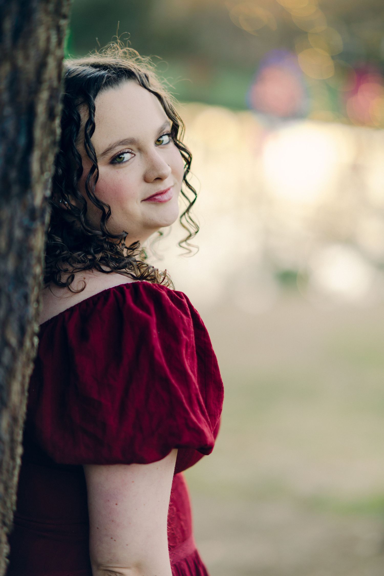 Chattanooga Senior girl with curly dark hair in a red velvet off-shoulder dress leaning against a tree, smiling softly outdoors.