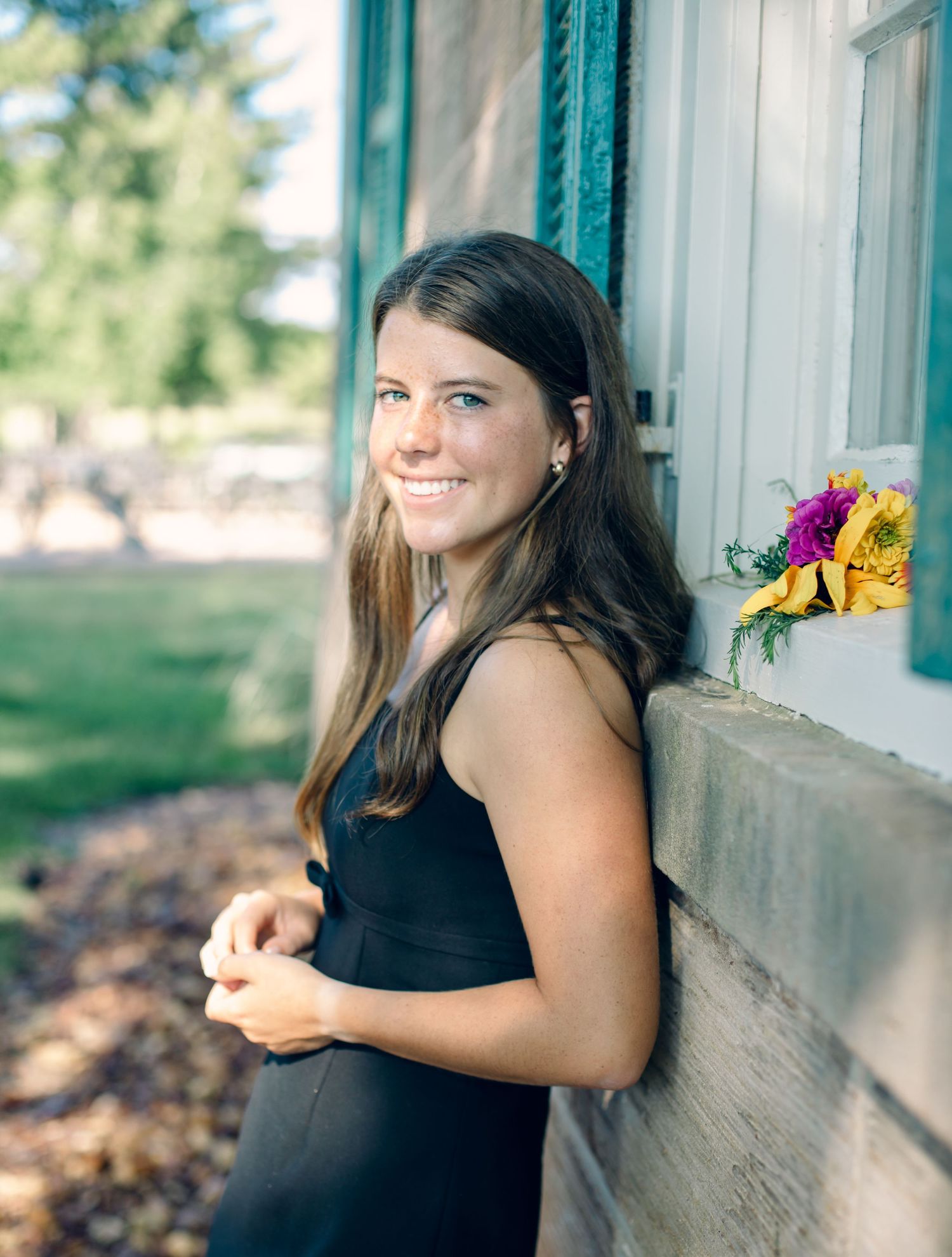 Girl in black dress smiling while leaning against white window frame with colorful flowers on windowsill outdoors.