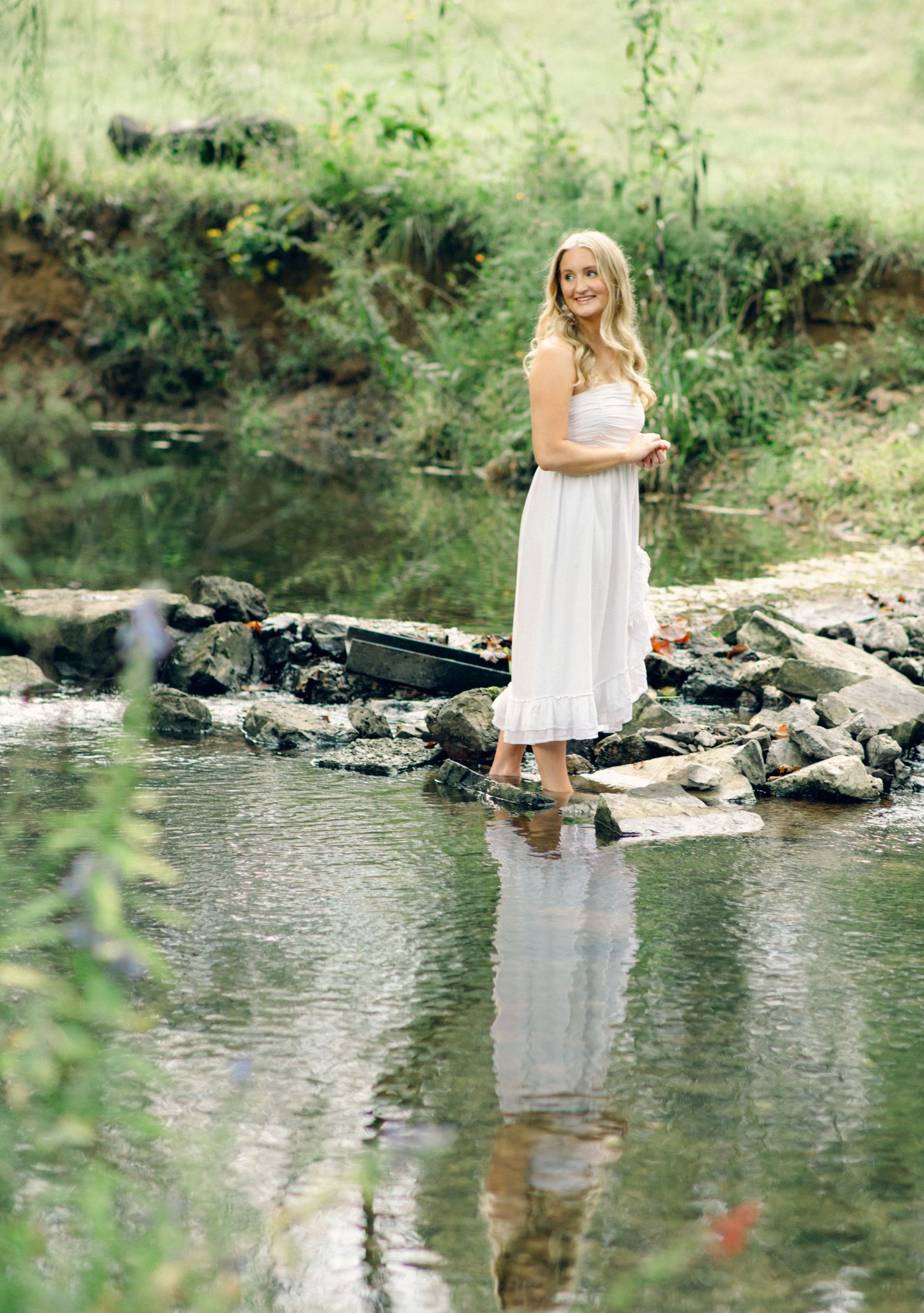 Blonde woman in white sundress standing on rocks in a serene creek surrounded by lush greenery.