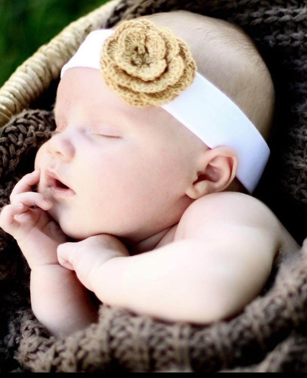 Sleeping newborn baby wearing a white headband with a tan crochet flower, nestled in a brown knit basket.