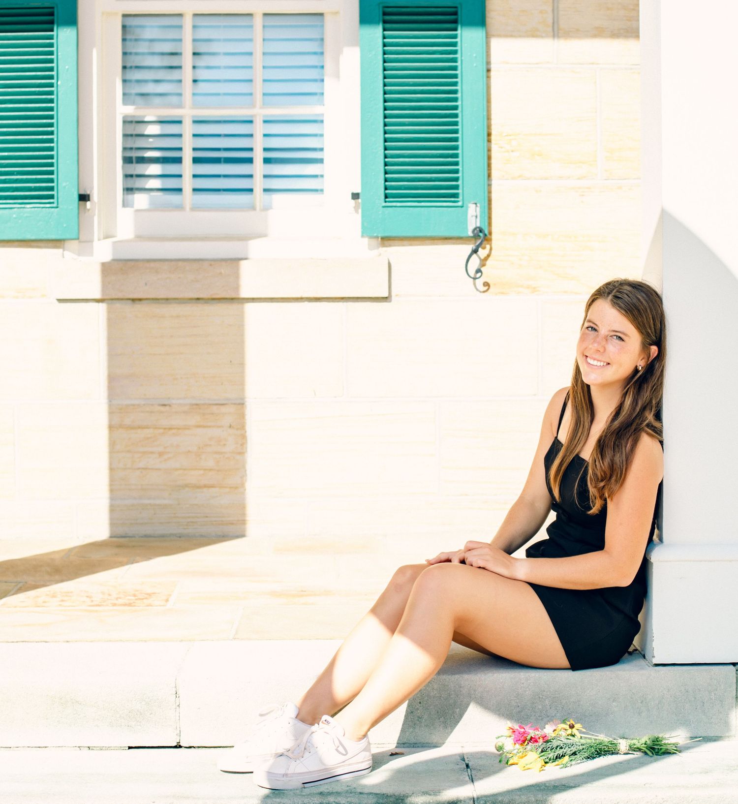 Young woman in black dress and white sneakers sitting on steps outside a building with teal shutters.