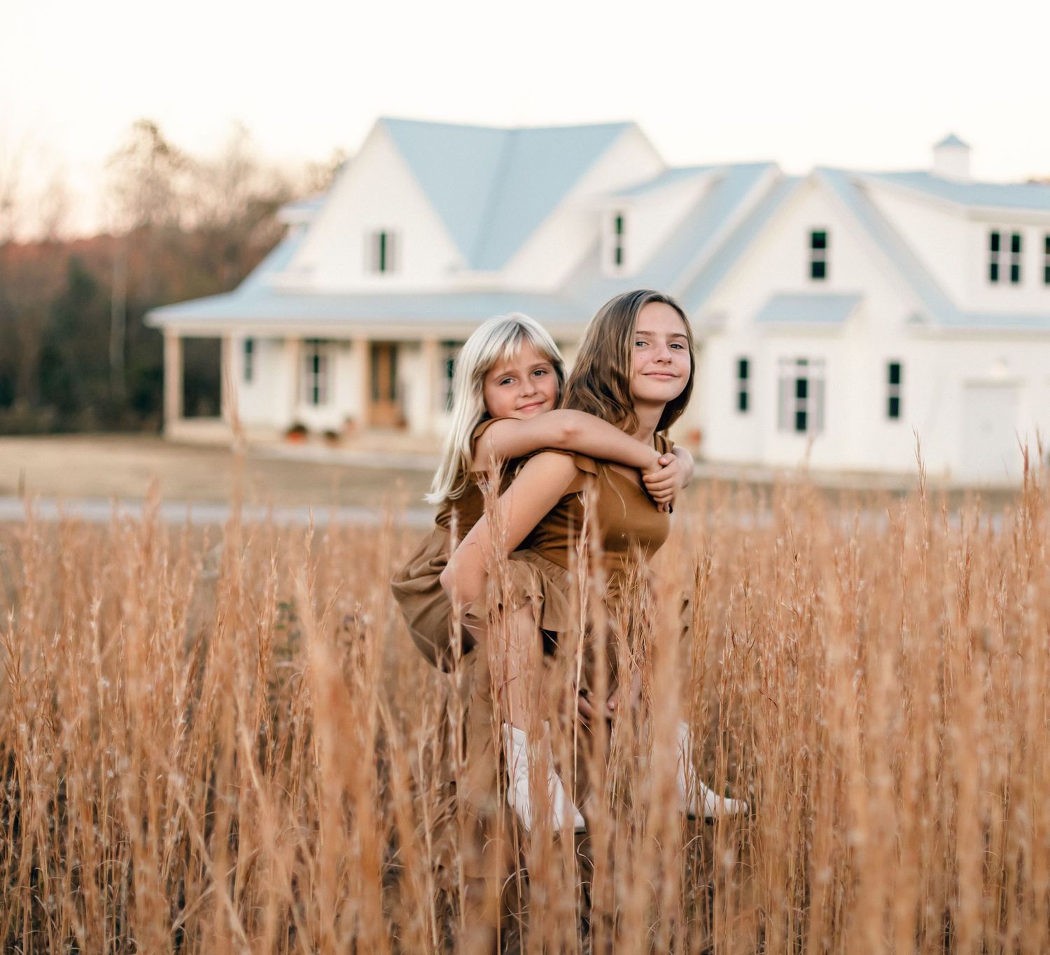 Two girls hugging in a golden field with a white farmhouse in the background during sunset.