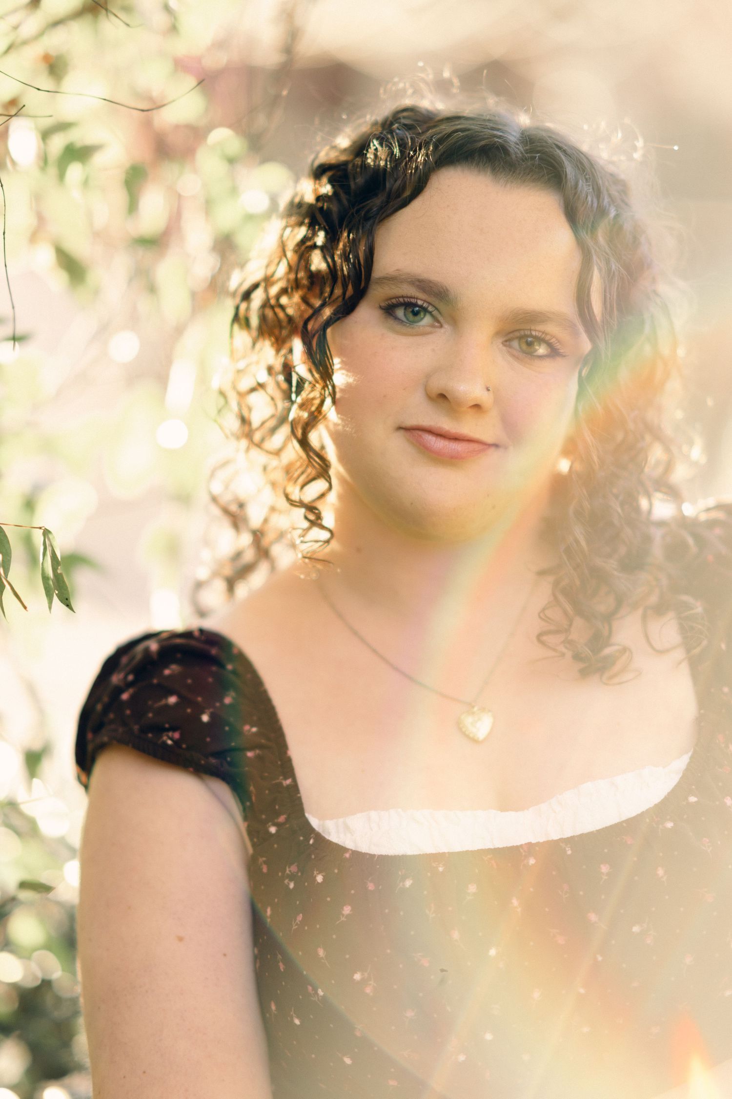 Young woman with curly hair wearing a heart necklace and dark dress, posing outdoors with soft bokeh lighting.