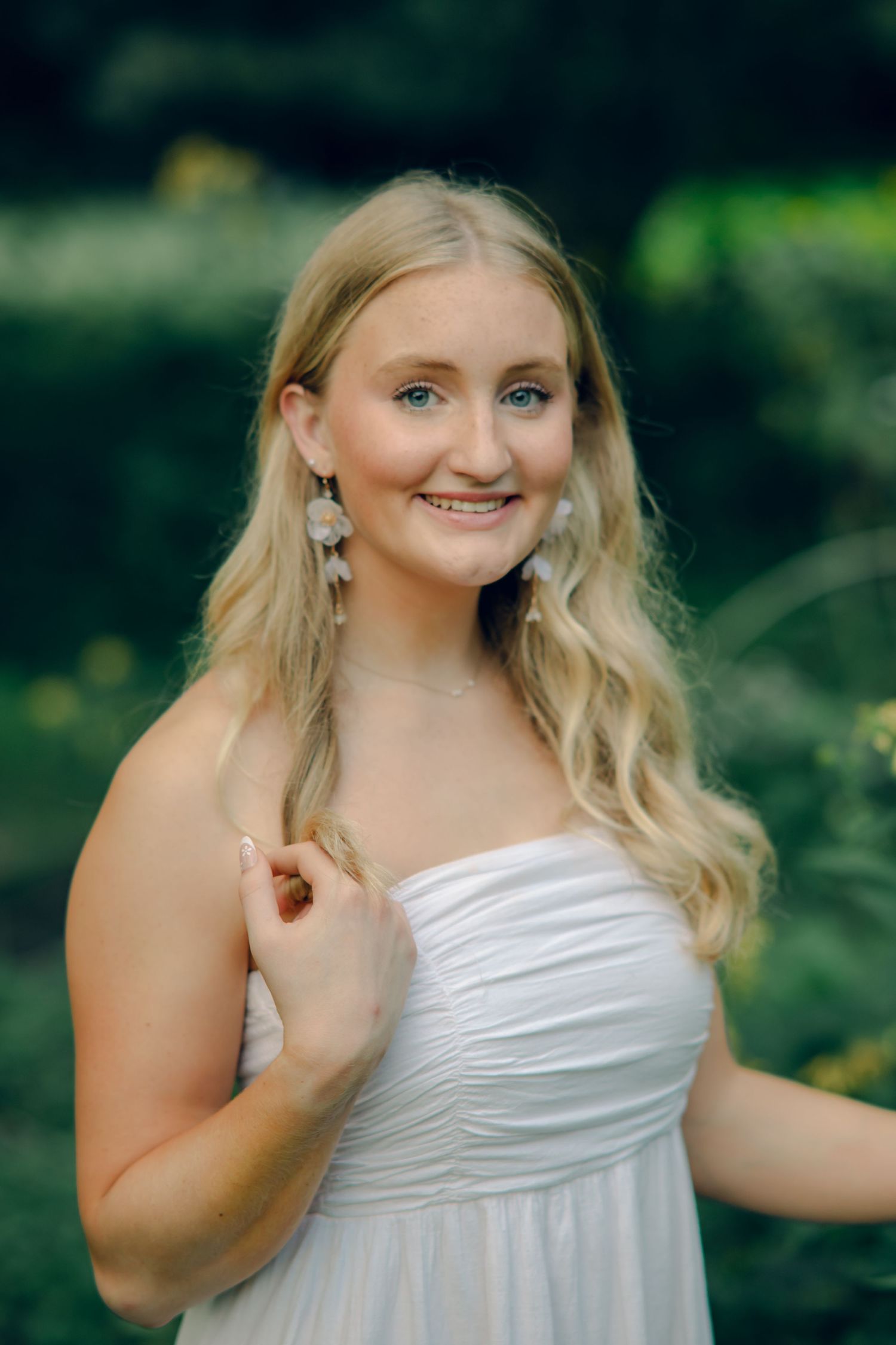 Young blonde woman in white strapless dress smiling outdoors with green foliage background.