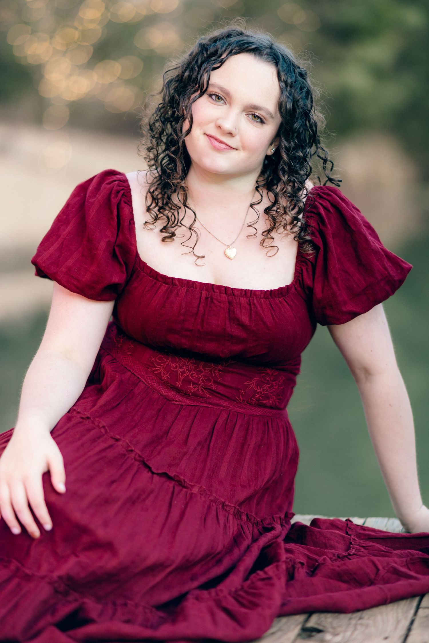Young woman with curly dark hair wearing a elegant crimson red dress, seated outdoors for a portrait photo.