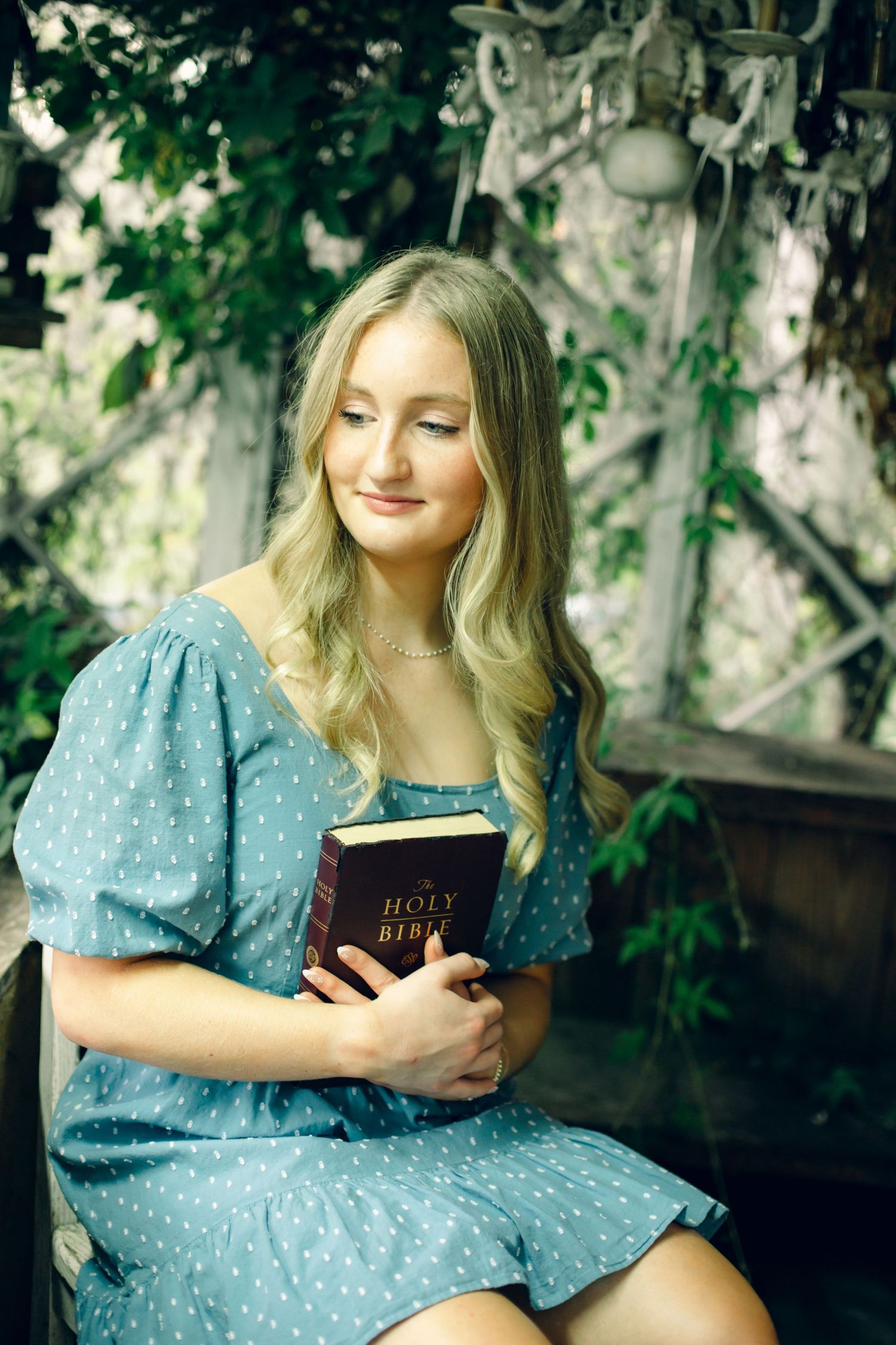Young blonde woman in blue polka dot dress holding a Holy Bible while sitting outdoors among green foliage.