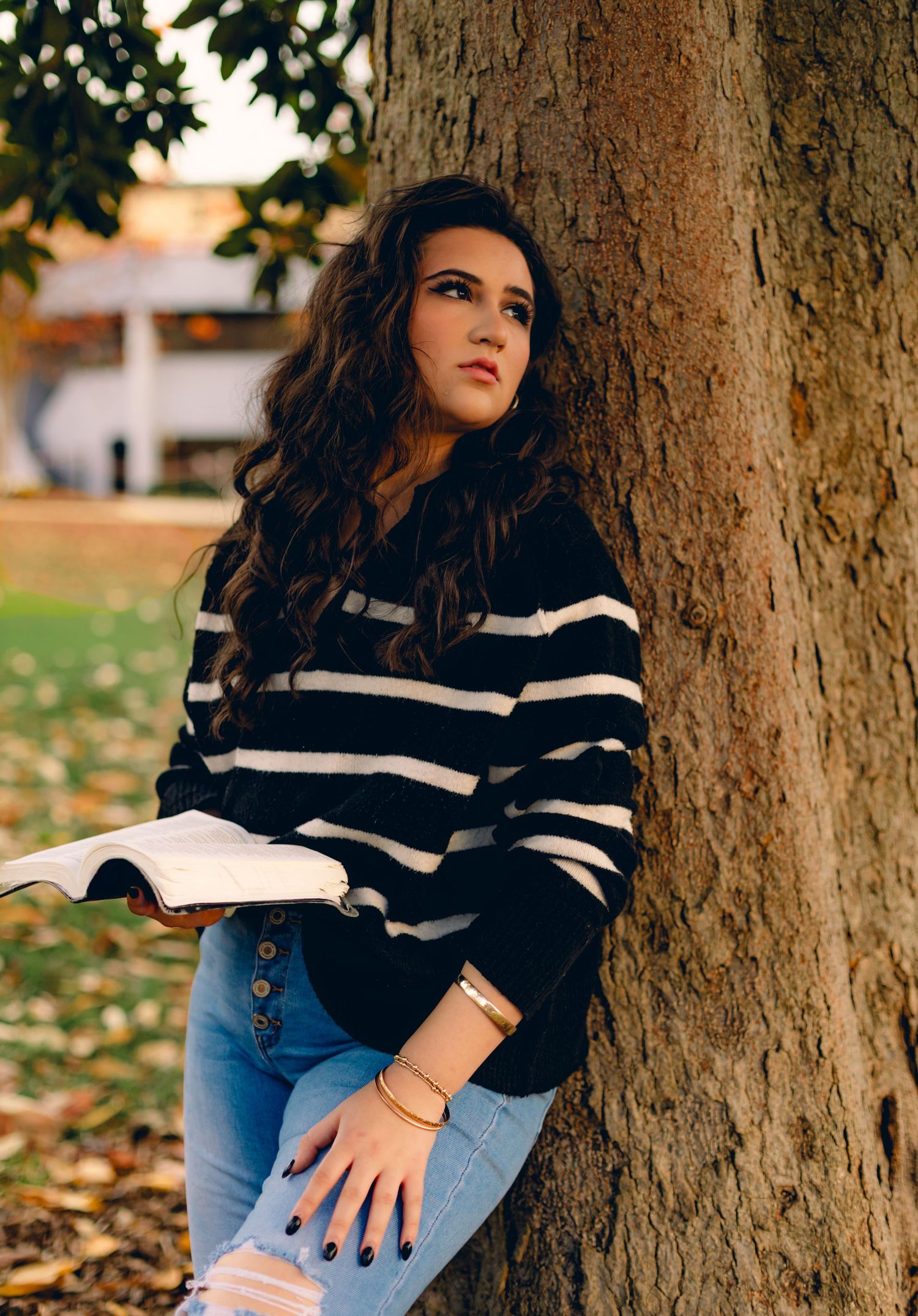 Young woman in striped sweater and jeans leaning against a tree, holding a book, gazing upward outdoors.