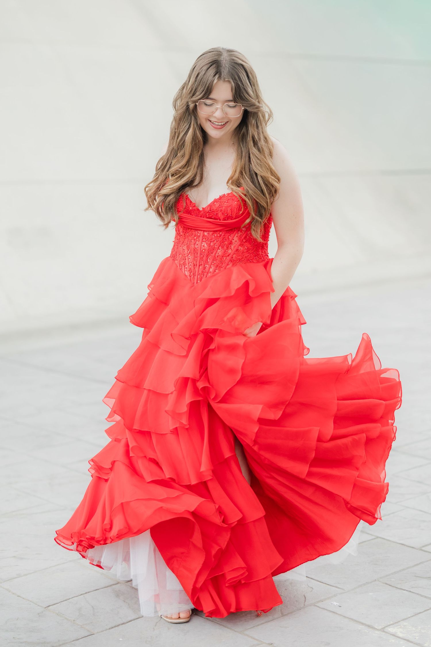 Young woman in a stunning red ruffled strapless ball gown twirling outdoors, with flowing layers and lace bodice.