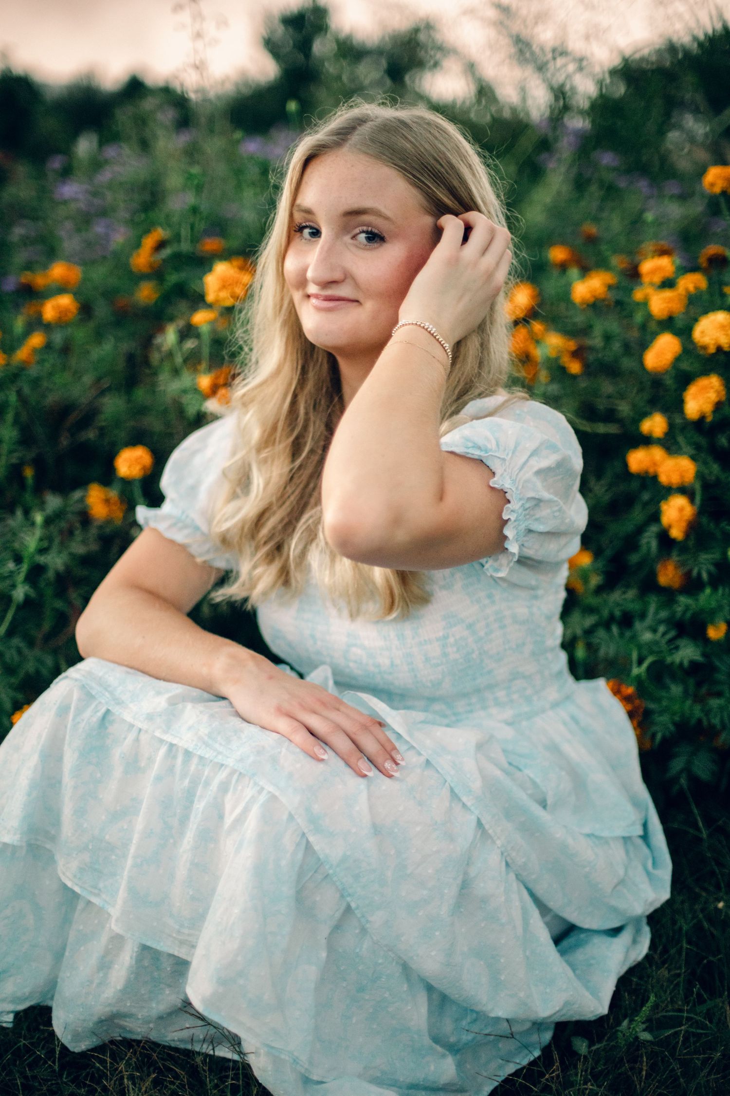 Young blonde woman in light blue dress sitting among orange marigolds in a garden at golden hour.