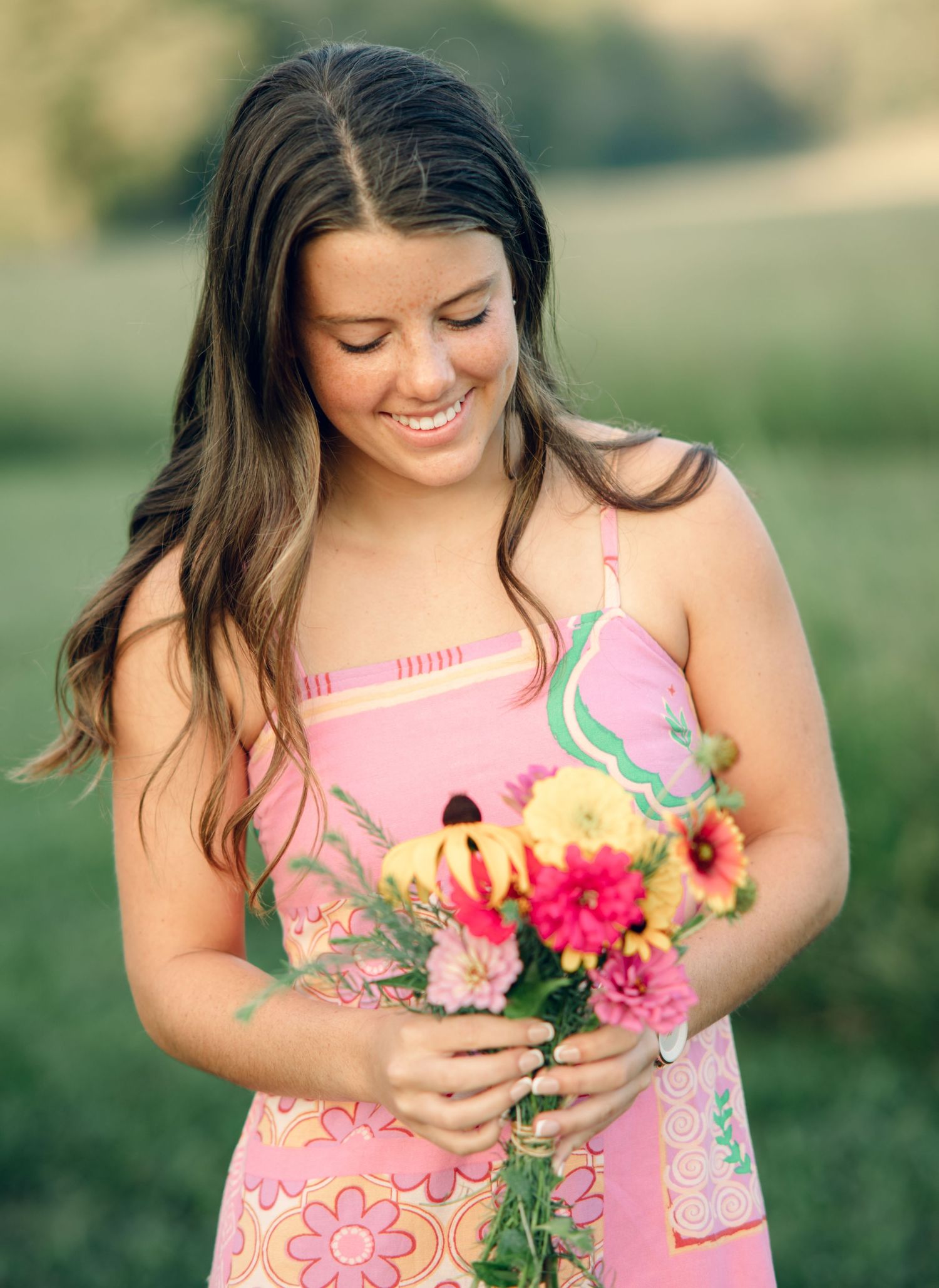 Young woman in pink floral dress smiling while holding a colorful bouquet of flowers outdoors in a green field.
