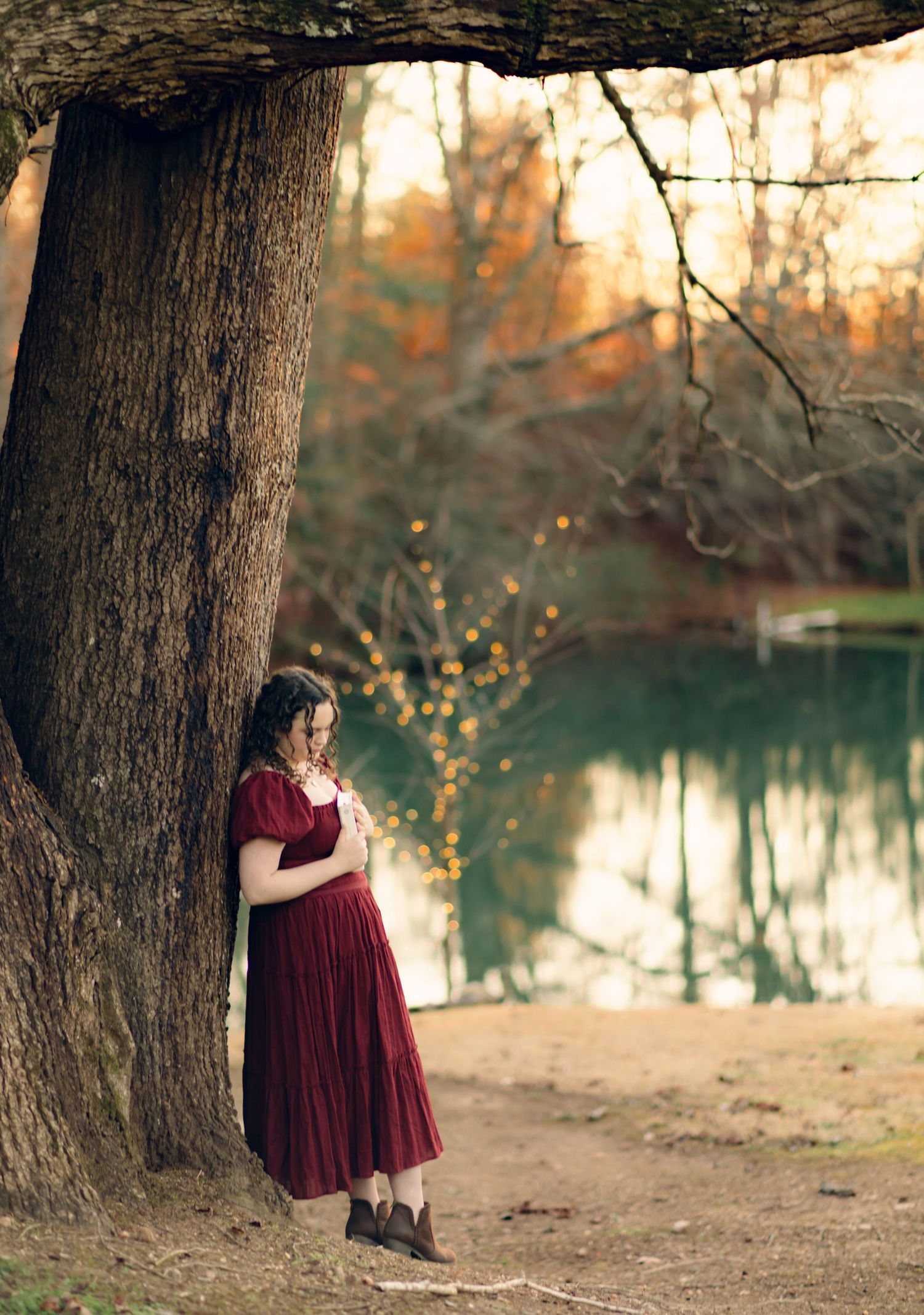 High school senior, Ashlyn in a red dress leaning against a large tree by a serene pond in autumn forest.