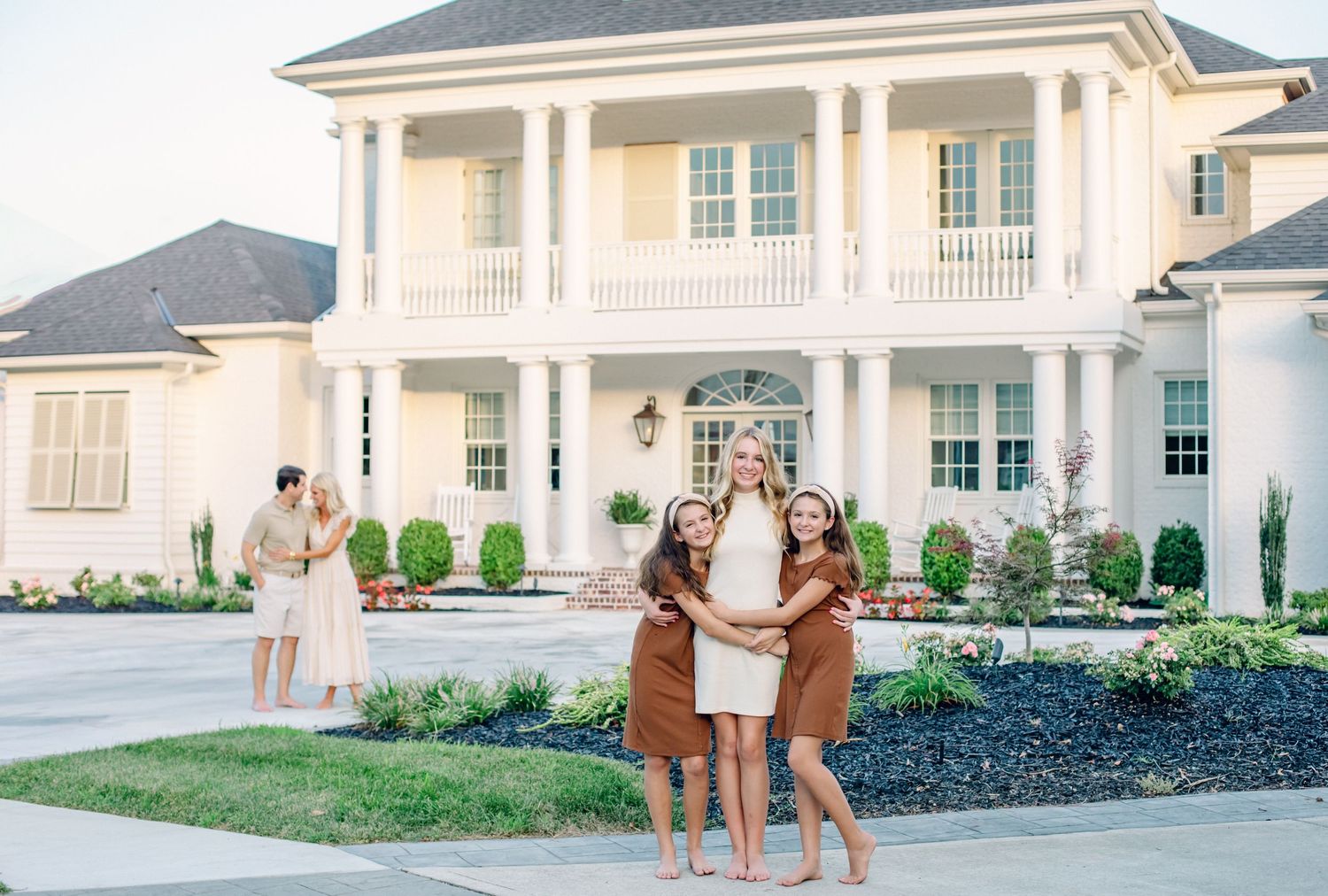 Family portrait of sisters hugging and smiling in front of a large white colonial-style home with manicured landscaping.