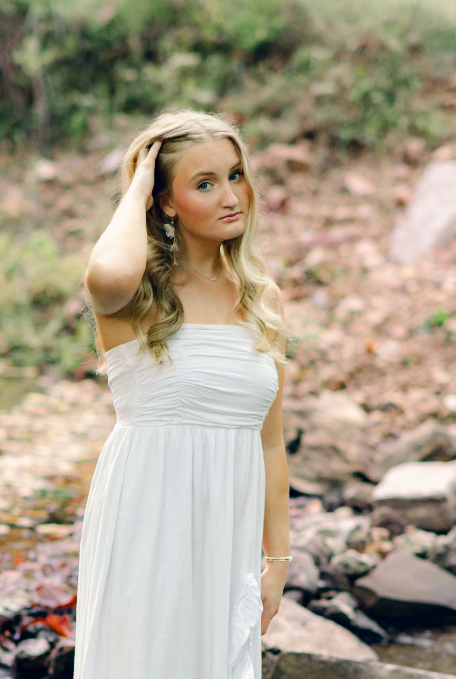 Young woman in white dress poses near rocky creek, hand in hair, with natural outdoor backdrop.