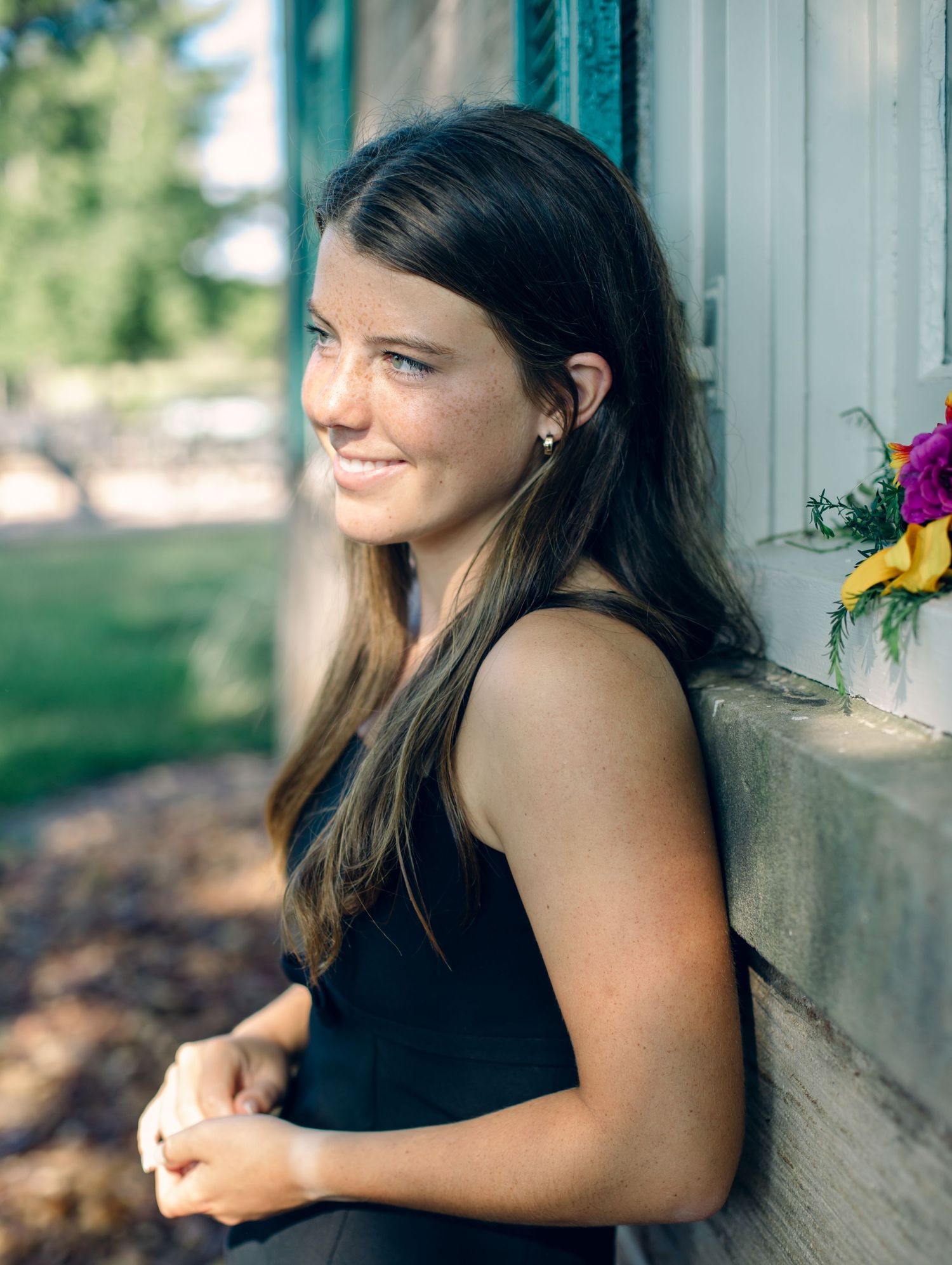Young woman with long brown hair wearing a black sleeveless top smiles while leaning against a white column outdoors.
