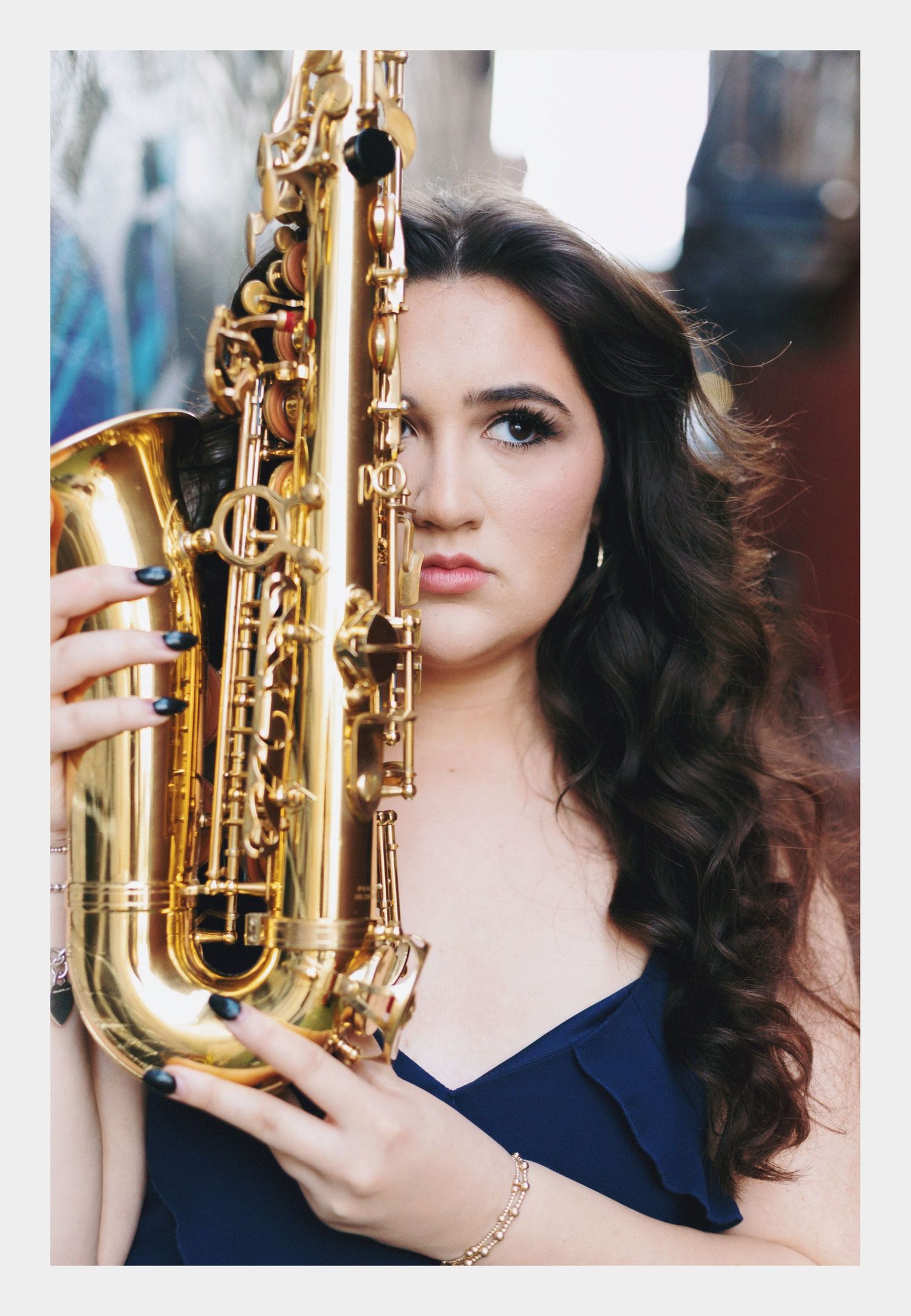 Young woman with dark curly hair holds a gold saxophone beside her face, wearing a navy blue dress.