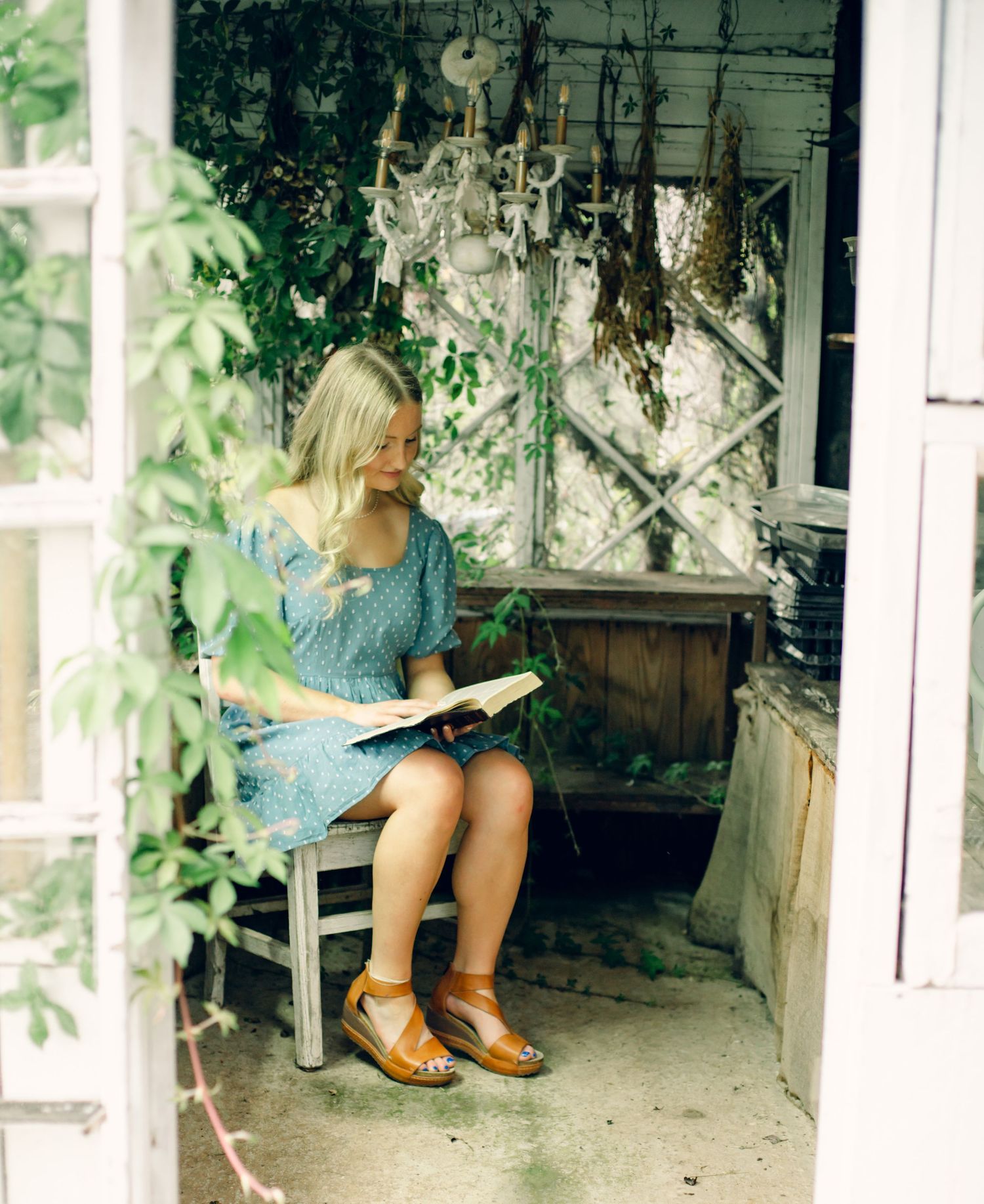Young blonde woman in blue dress reading a book on a bench in a charming rustic garden greenhouse.