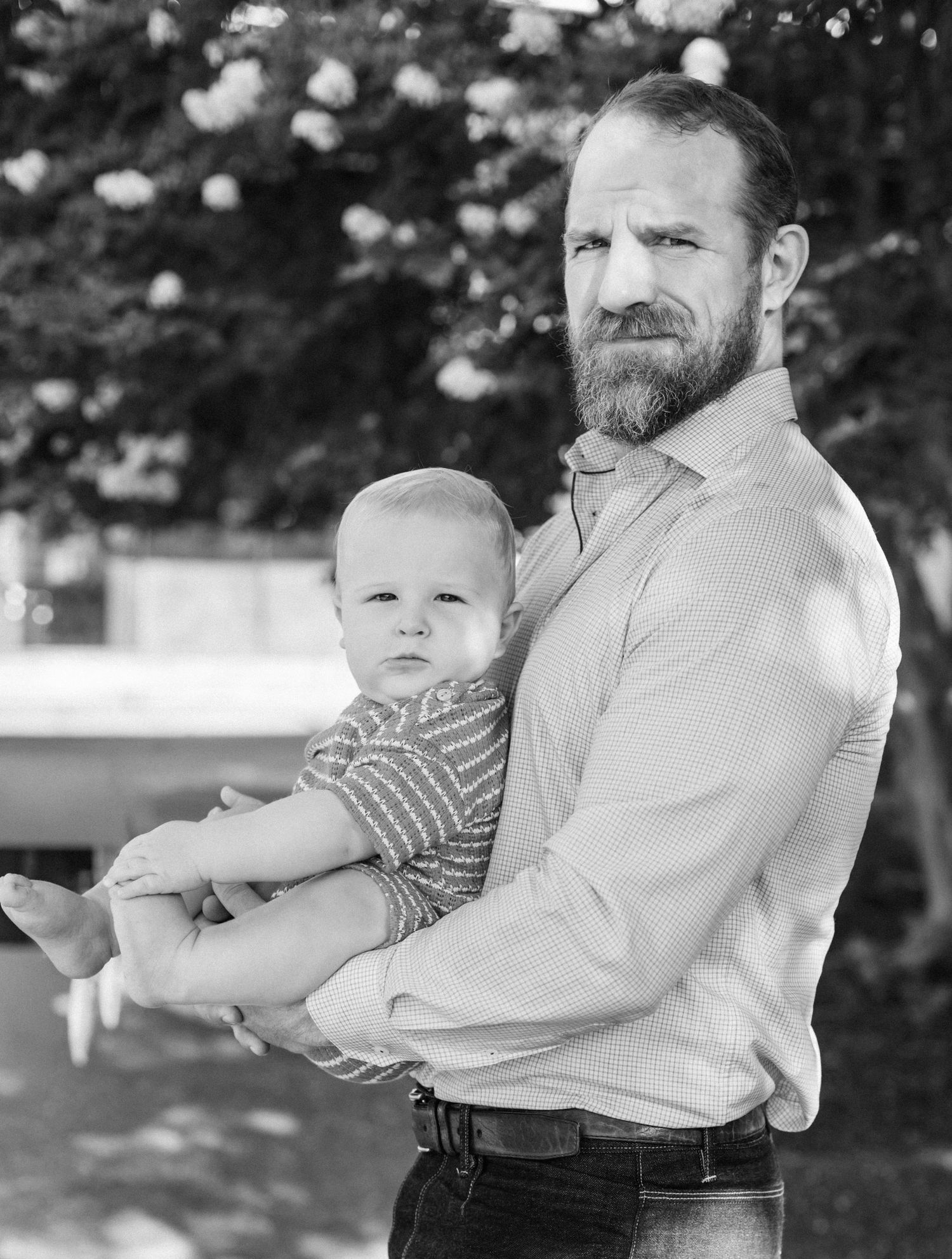 Black and white portrait of bearded man holding a baby outdoors with blurred trees in background.