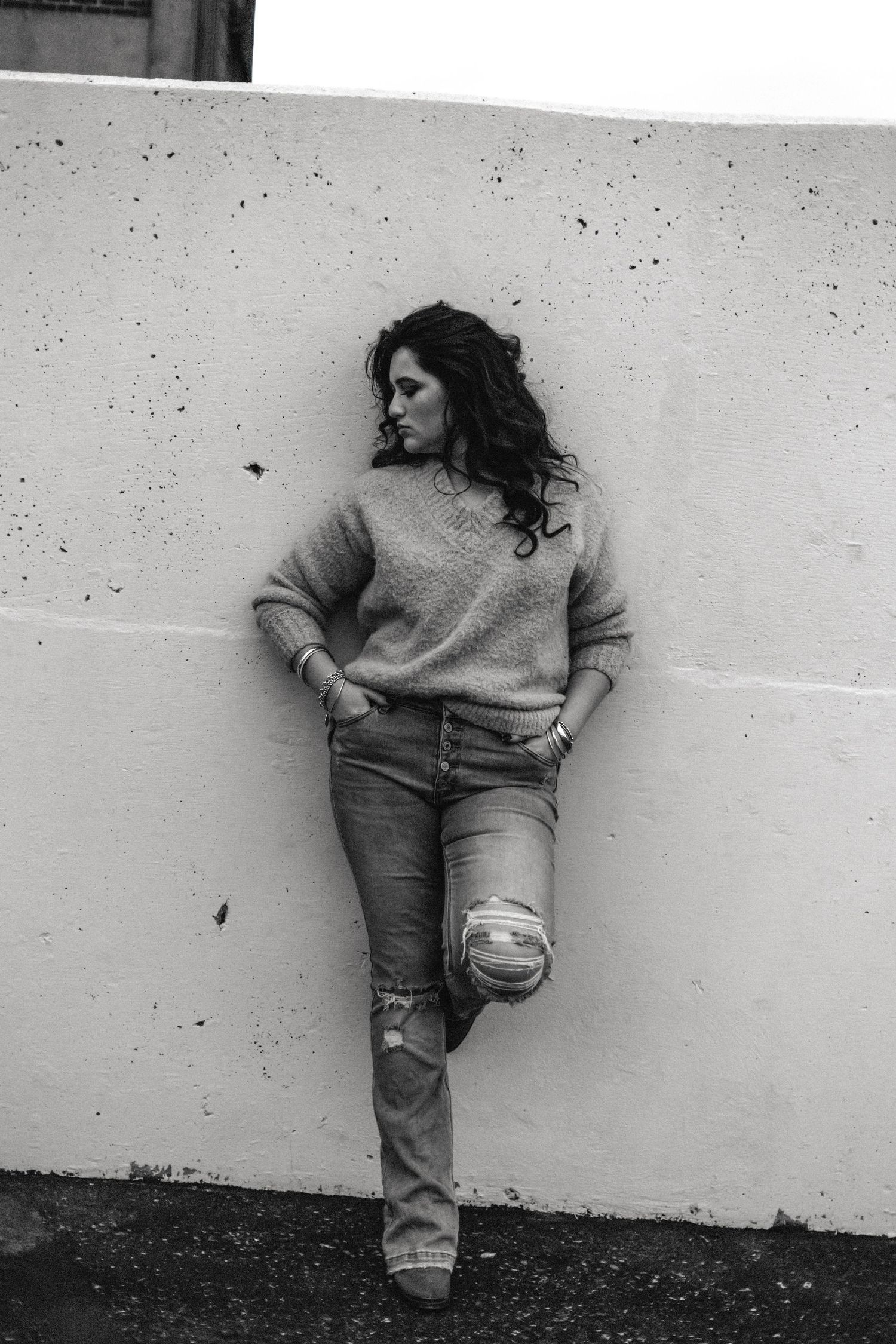 Black and white photo of a woman in ripped jeans and sweater leaning against a white wall outdoors.