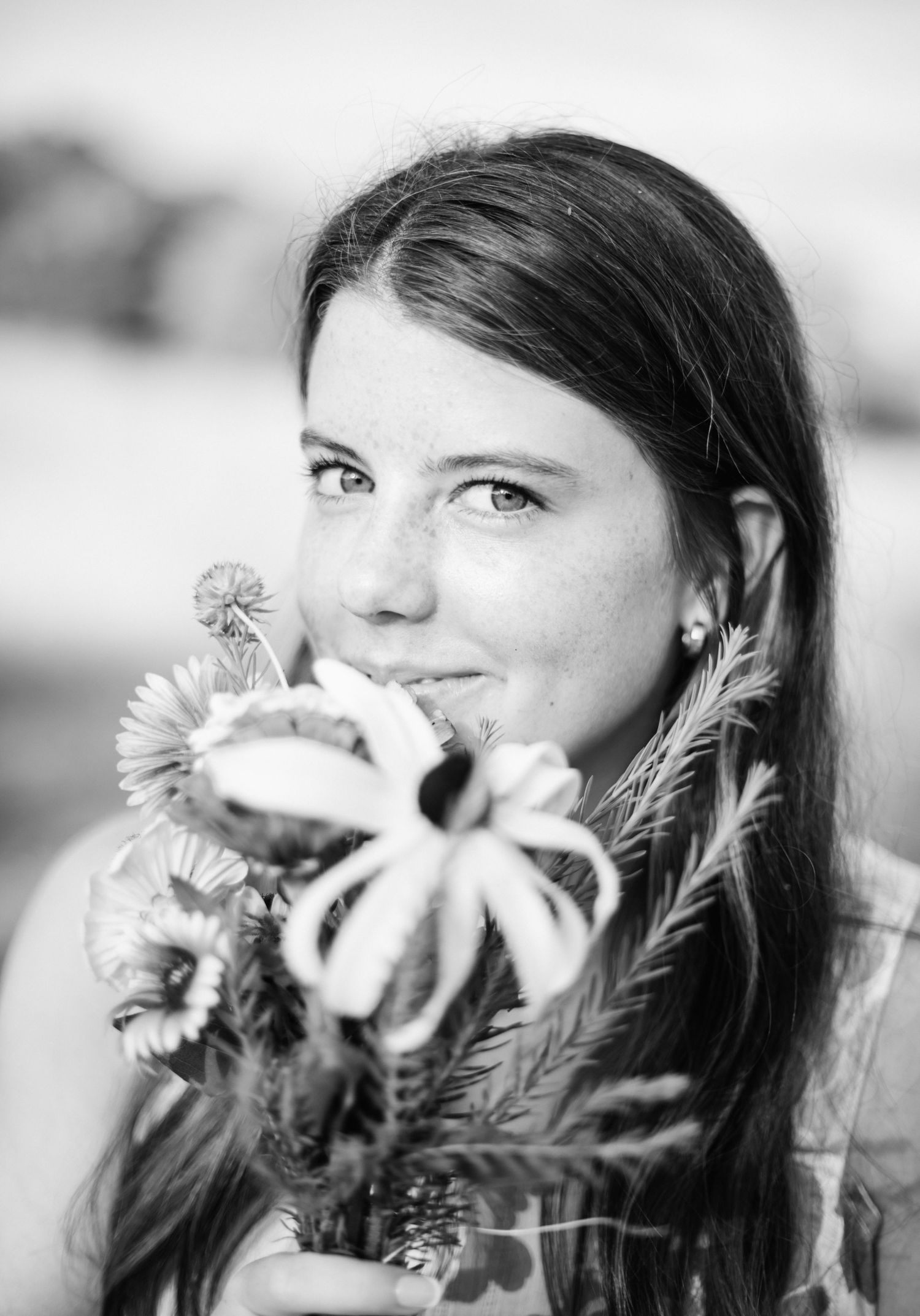Young woman with long dark hair holding wildflowers, smiling softly in a black and white portrait photo.