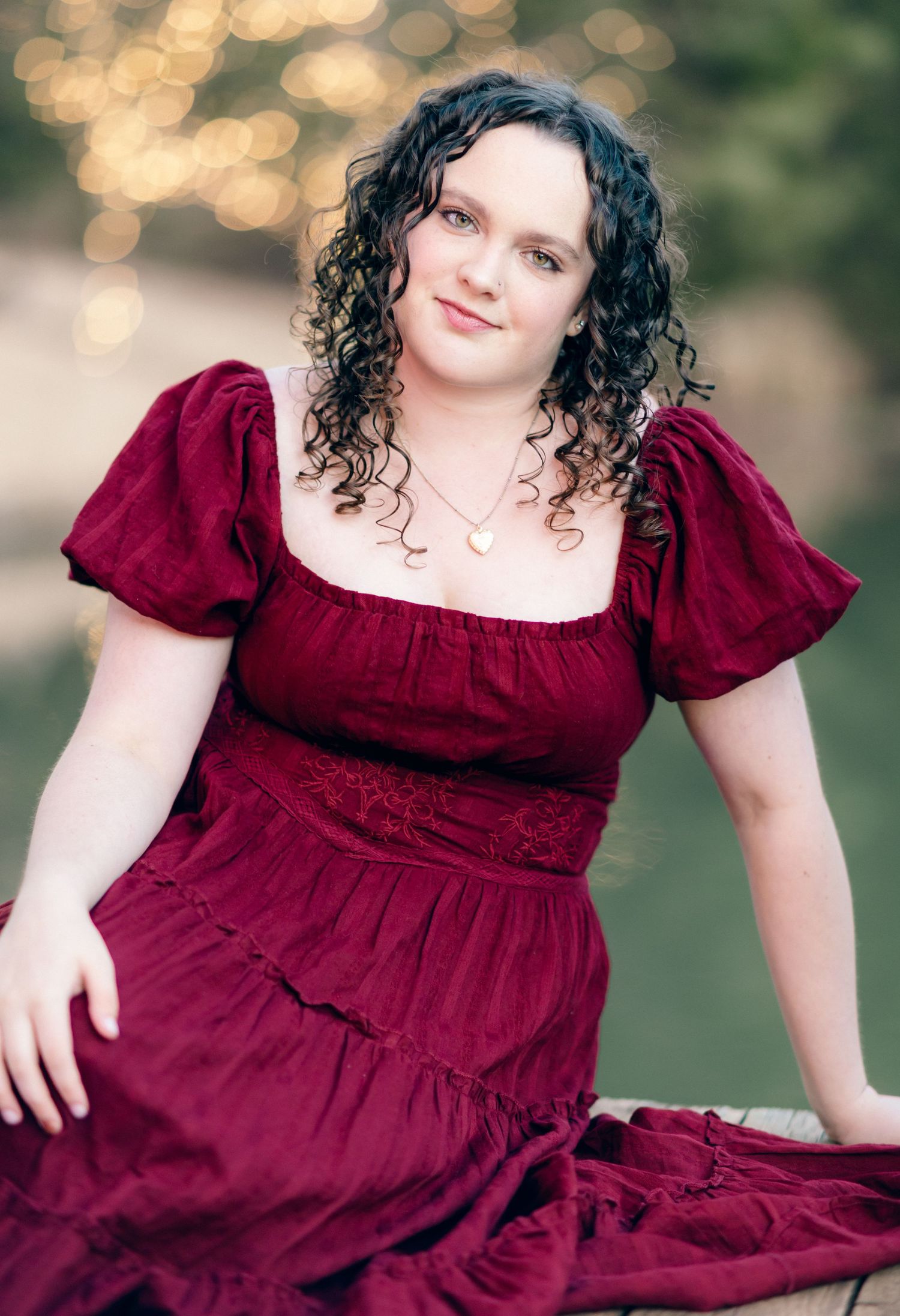 Young woman with curly dark hair wearing a crimson red puff-sleeve dress, posing outdoors with bokeh lighting.