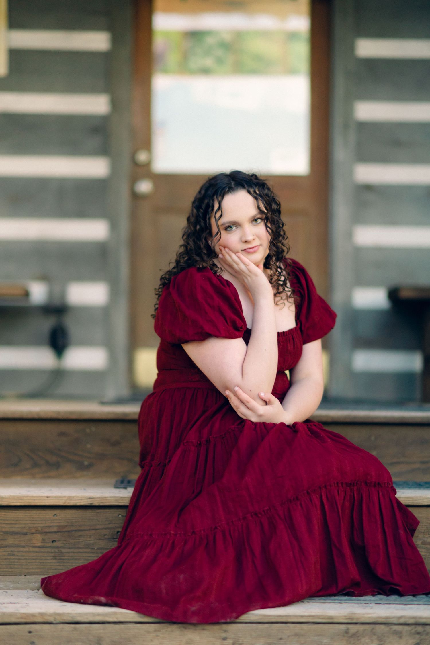 Young woman with curly dark hair in a red velvet dress sitting on wooden porch steps, posing with hand on chin.