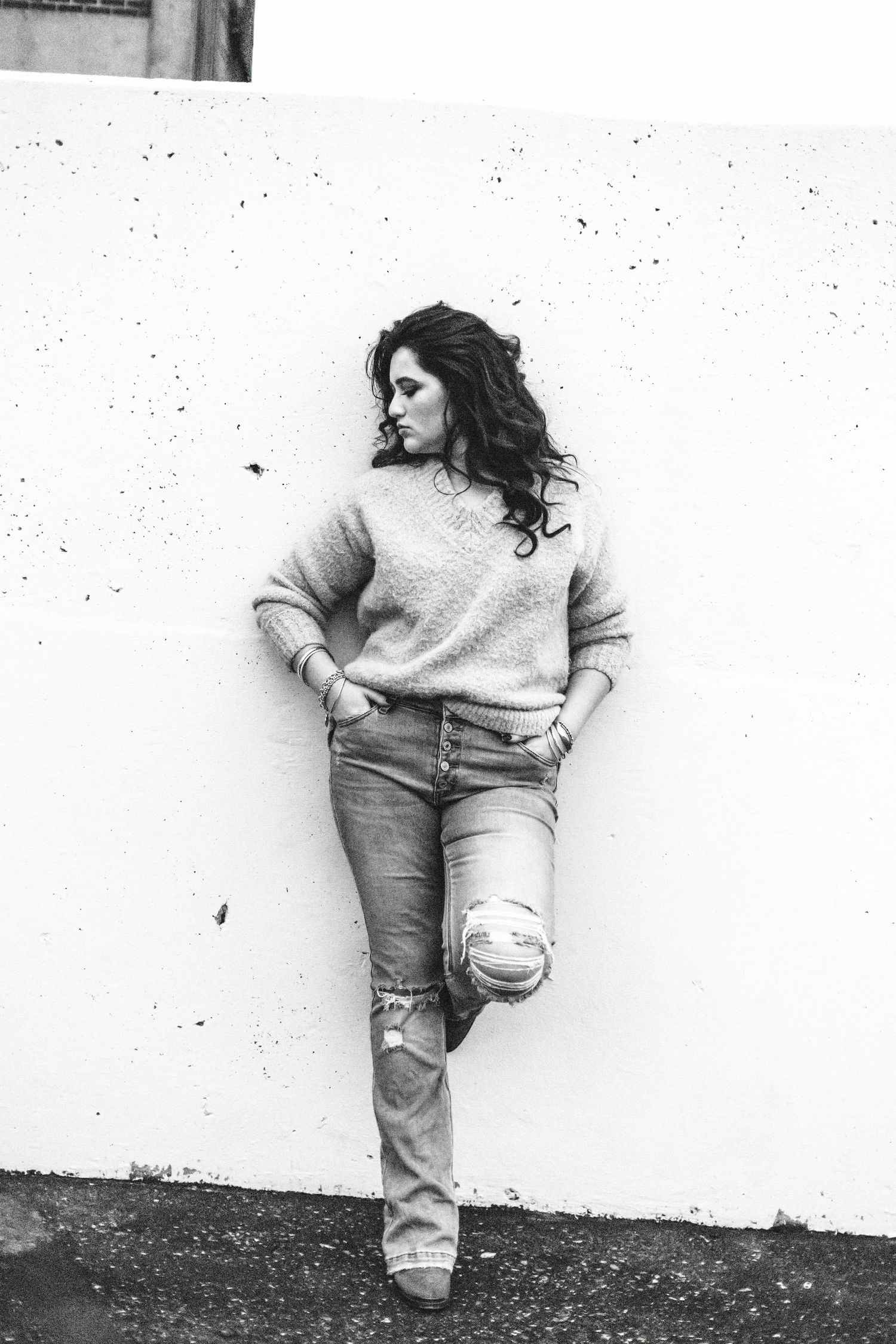 Black and white photo of a woman in jeans and sweater leaning against a white wall with one leg crossed.