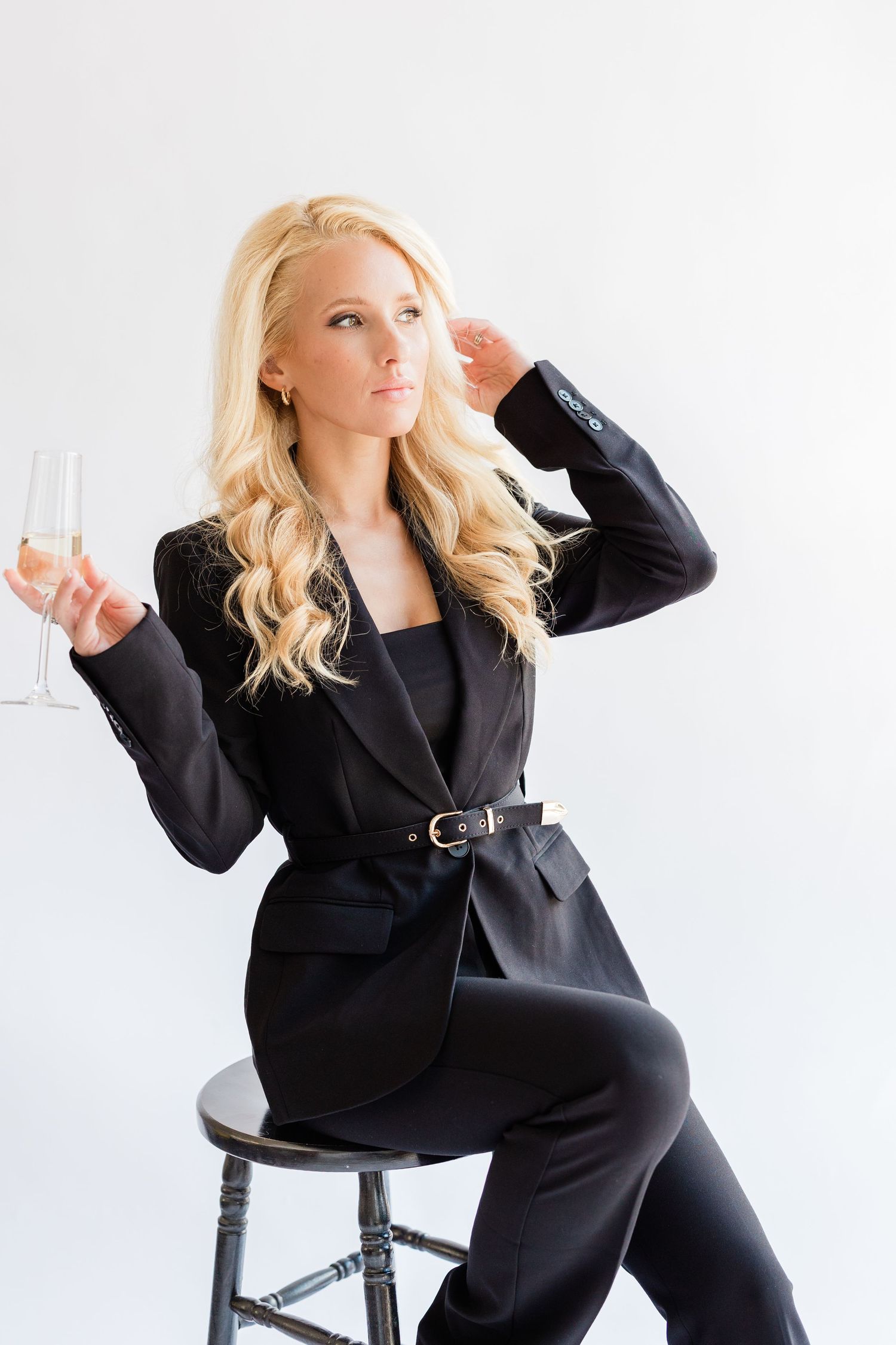 Blonde woman in black suit sitting on stool holding champagne glass against white background.