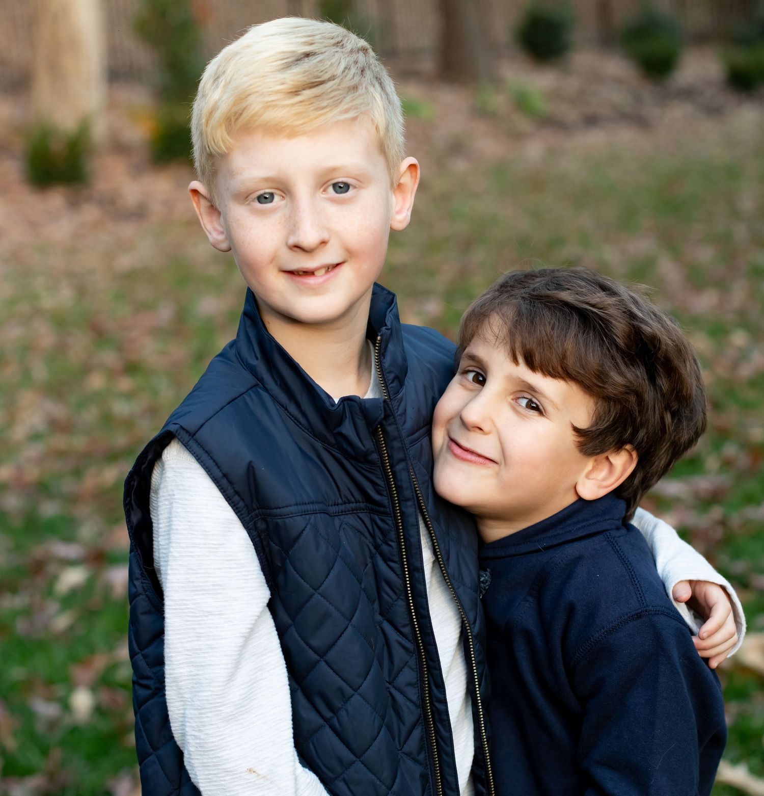 Two young boys in navy blue vests smiling and posing together outdoors in a park setting.
