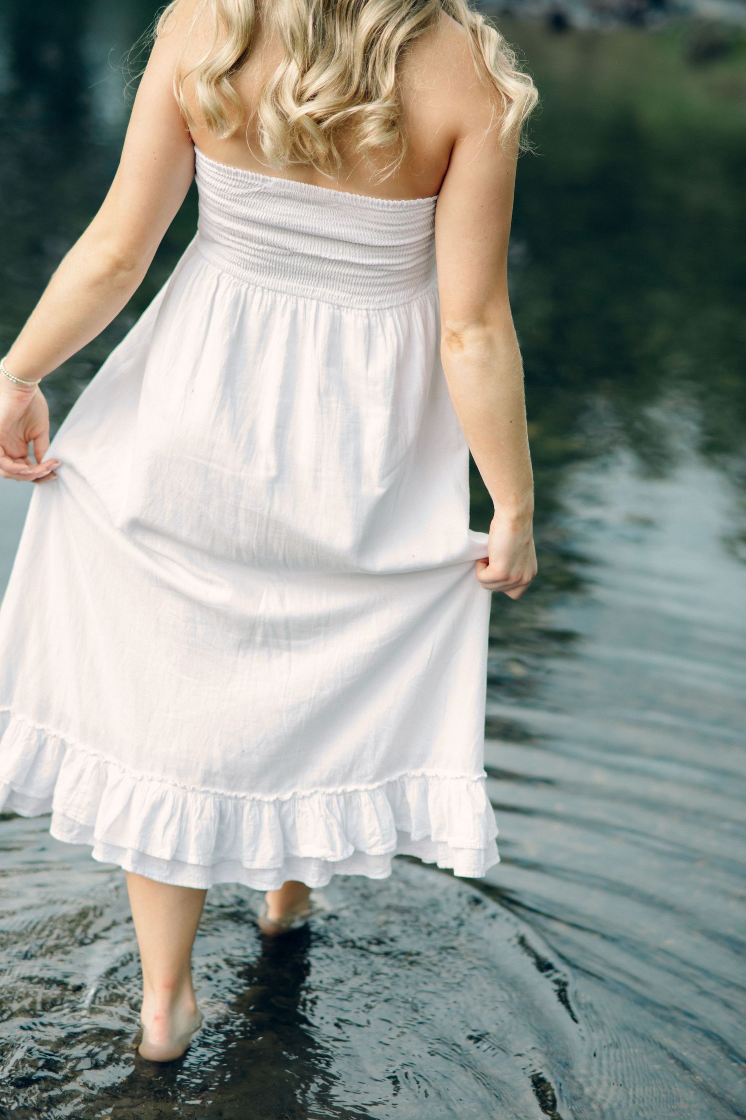 A blonde girl in white strapless maxi dress wading barefoot in calm water, viewed from behind.