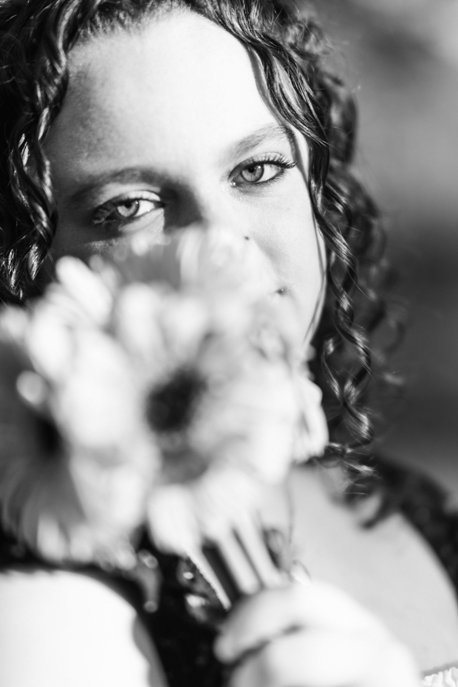 Black and white senior session portrait of Ashlyn with curly hair holding a flower in front of her face.
