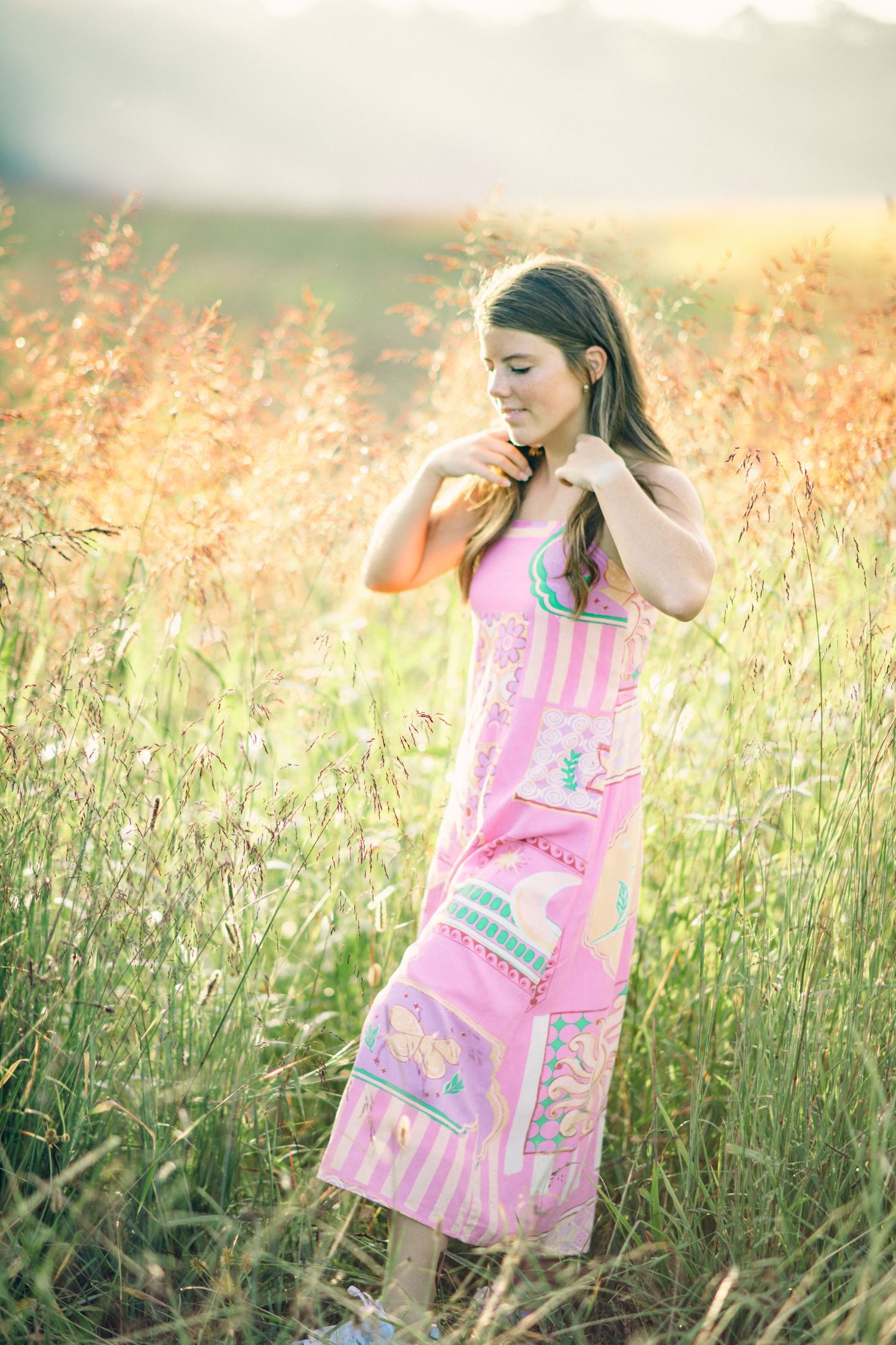 Young woman in pink maxi dress standing in golden field of tall grass during sunset.