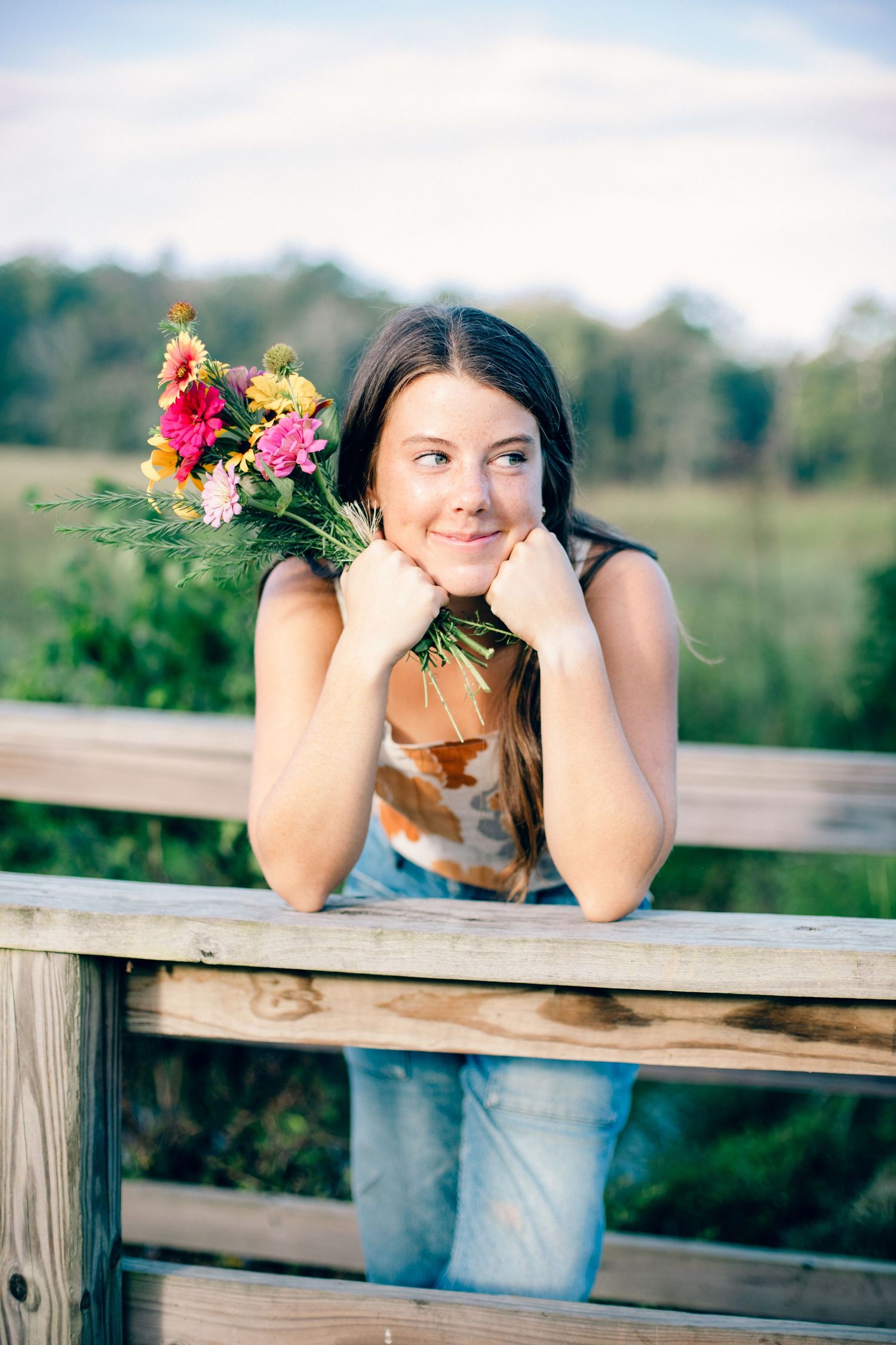 Senior Eden leaning on wooden fence holding colorful wildflower bouquet, smiling outdoors in a green field. At Chickamauga Battlefield.