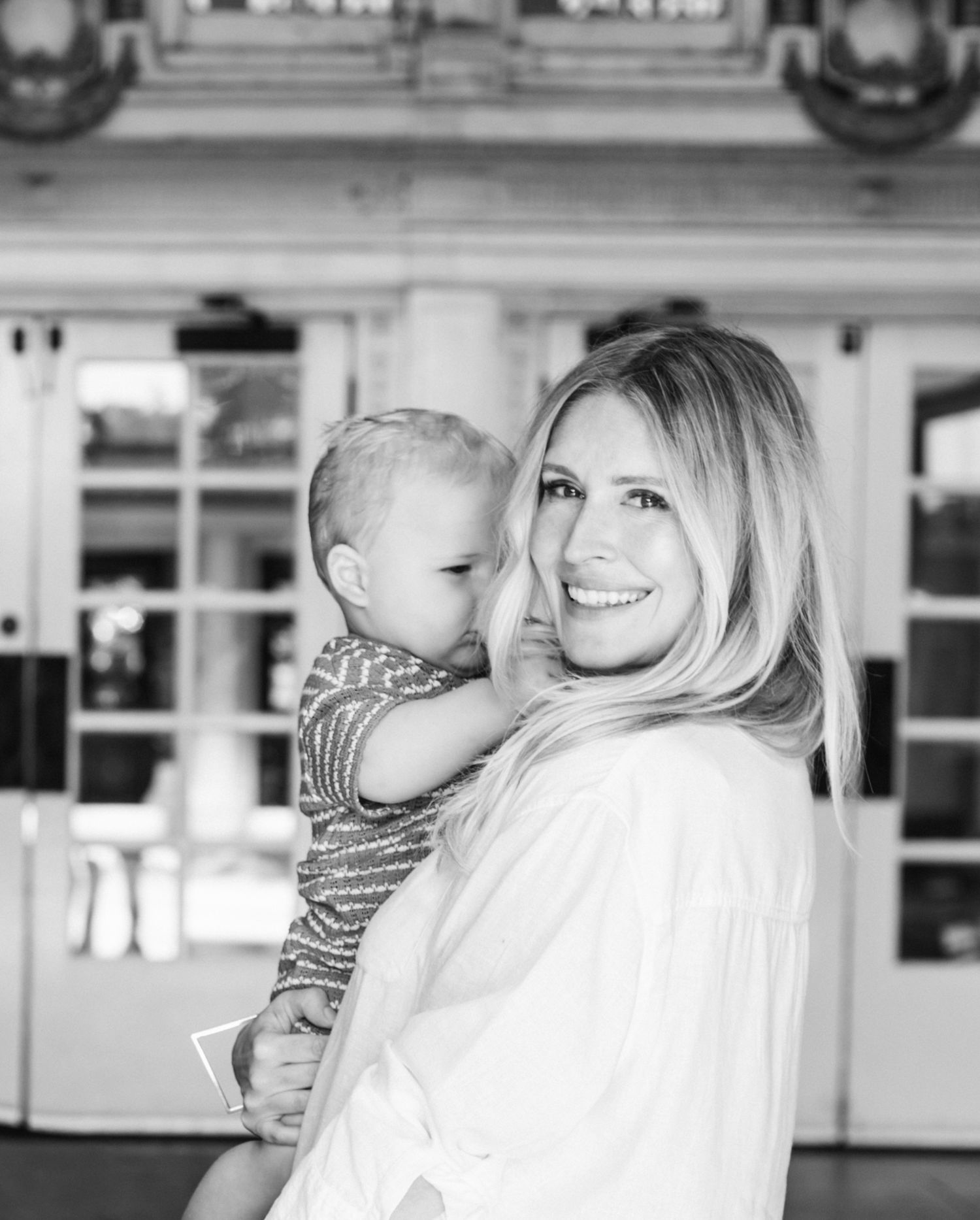 Black and white photo of a smiling woman with long blonde hair holding a young toddler outdoors.