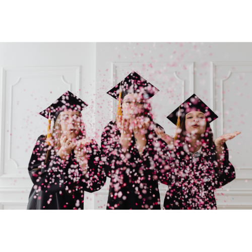 Three graduates in black caps and gowns celebrate by blowing pink confetti into the air indoors.