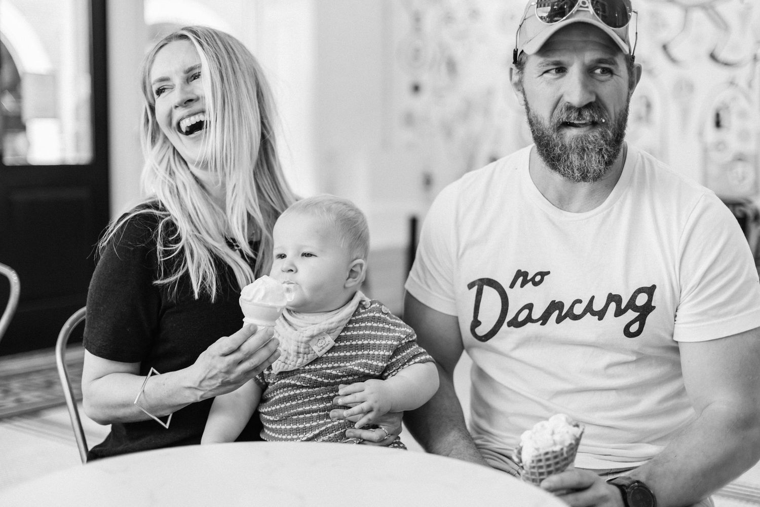 Black and white photo of a smiling woman, baby, and bearded man in a No Dancing shirt sitting together.
