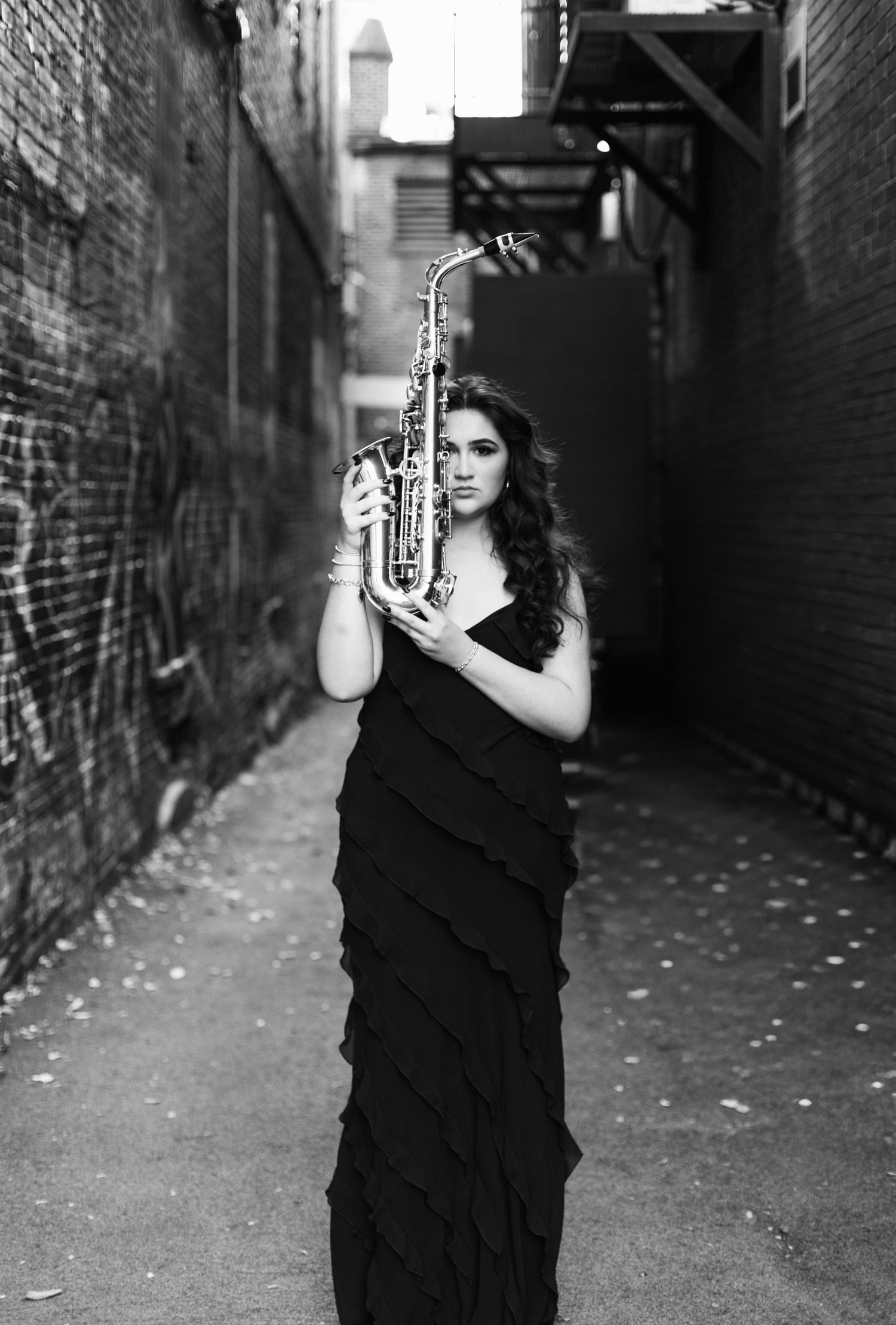 Woman in black gown holding saxophone in urban alley, black and white portrait photography.