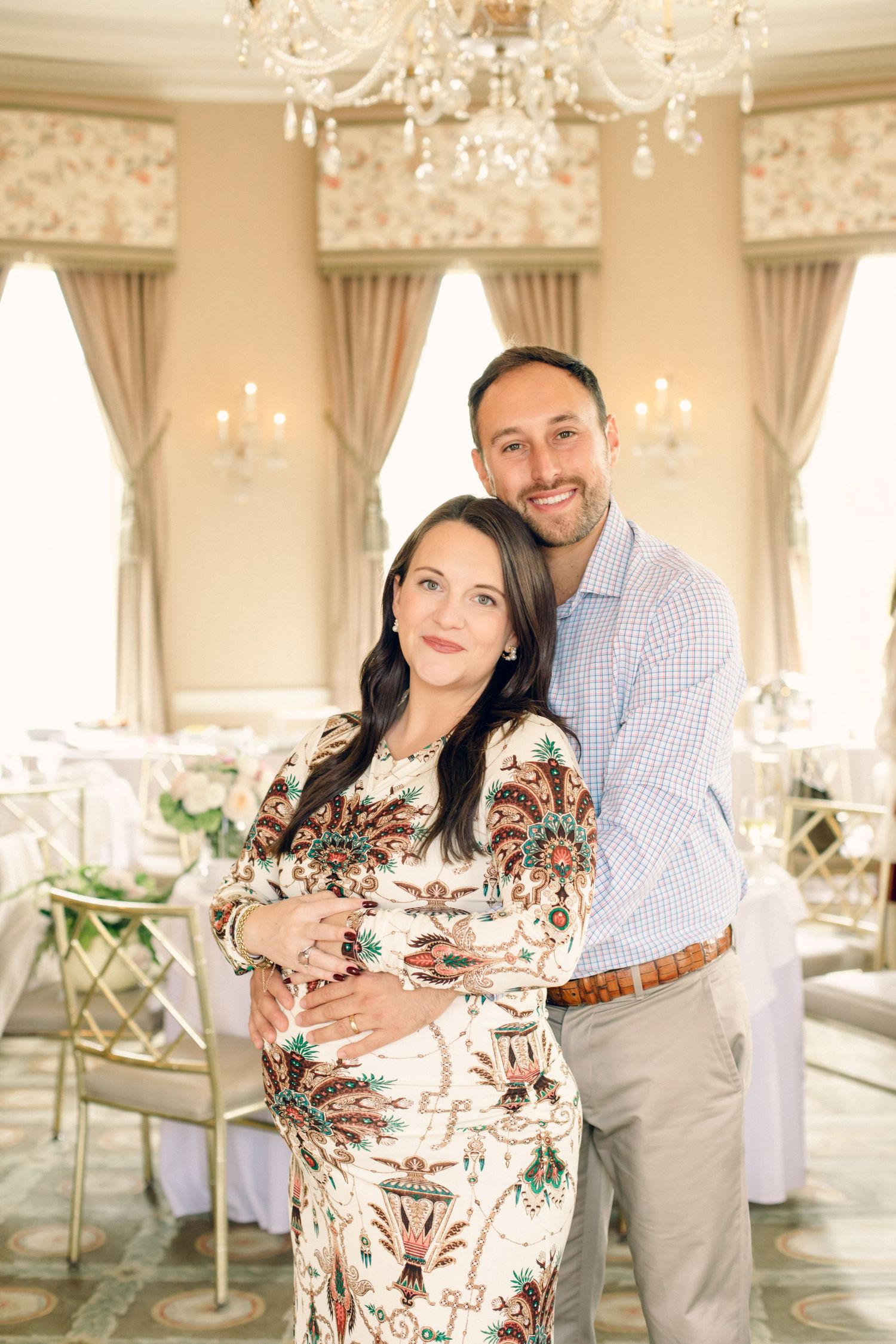 Pregnant couple posing together in elegant ballroom with chandeliers, woman in floral dress, man in light blue shirt.