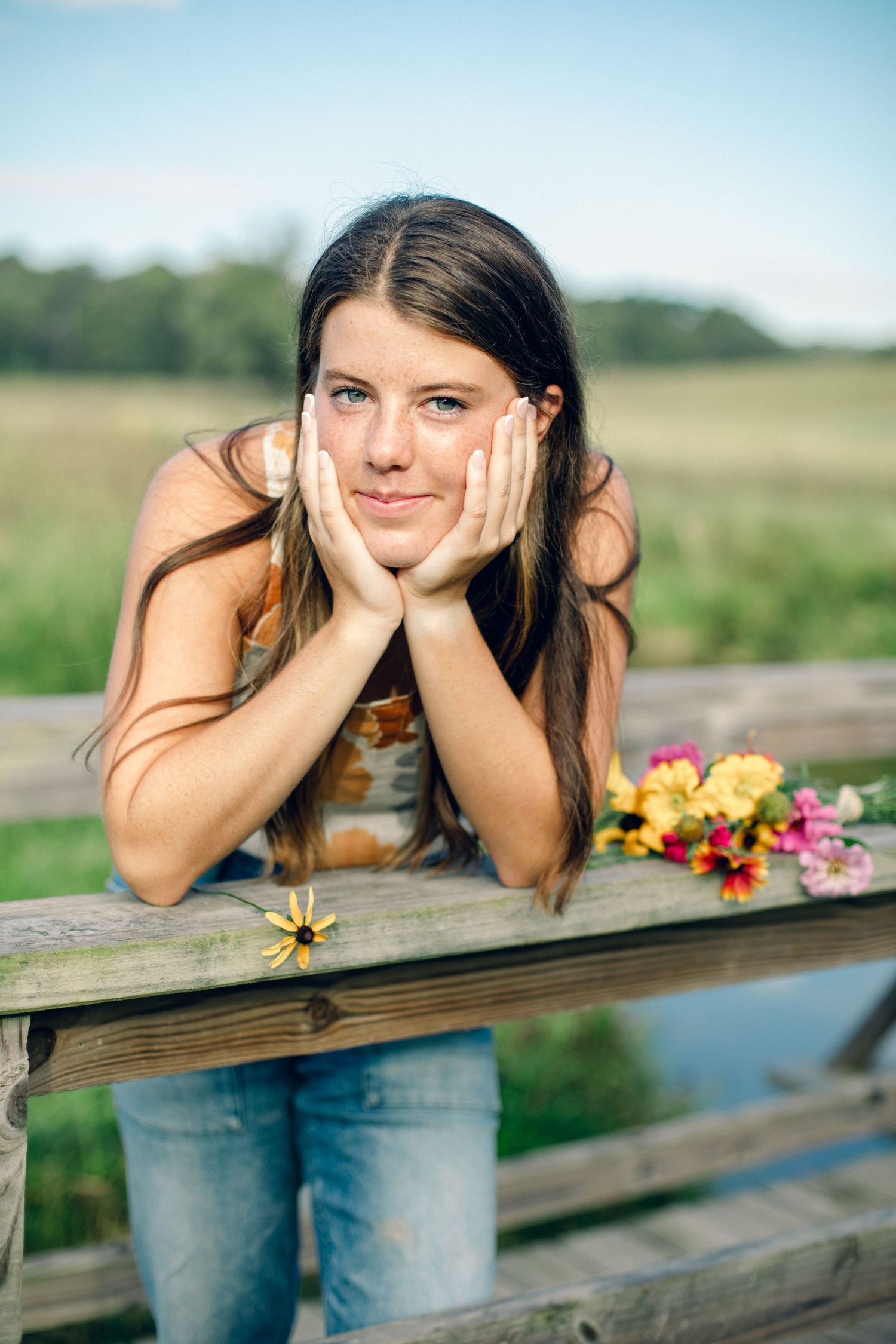 Young woman with long brown hair resting chin on hands on wooden fence, with colorful wildflowers in a green field.