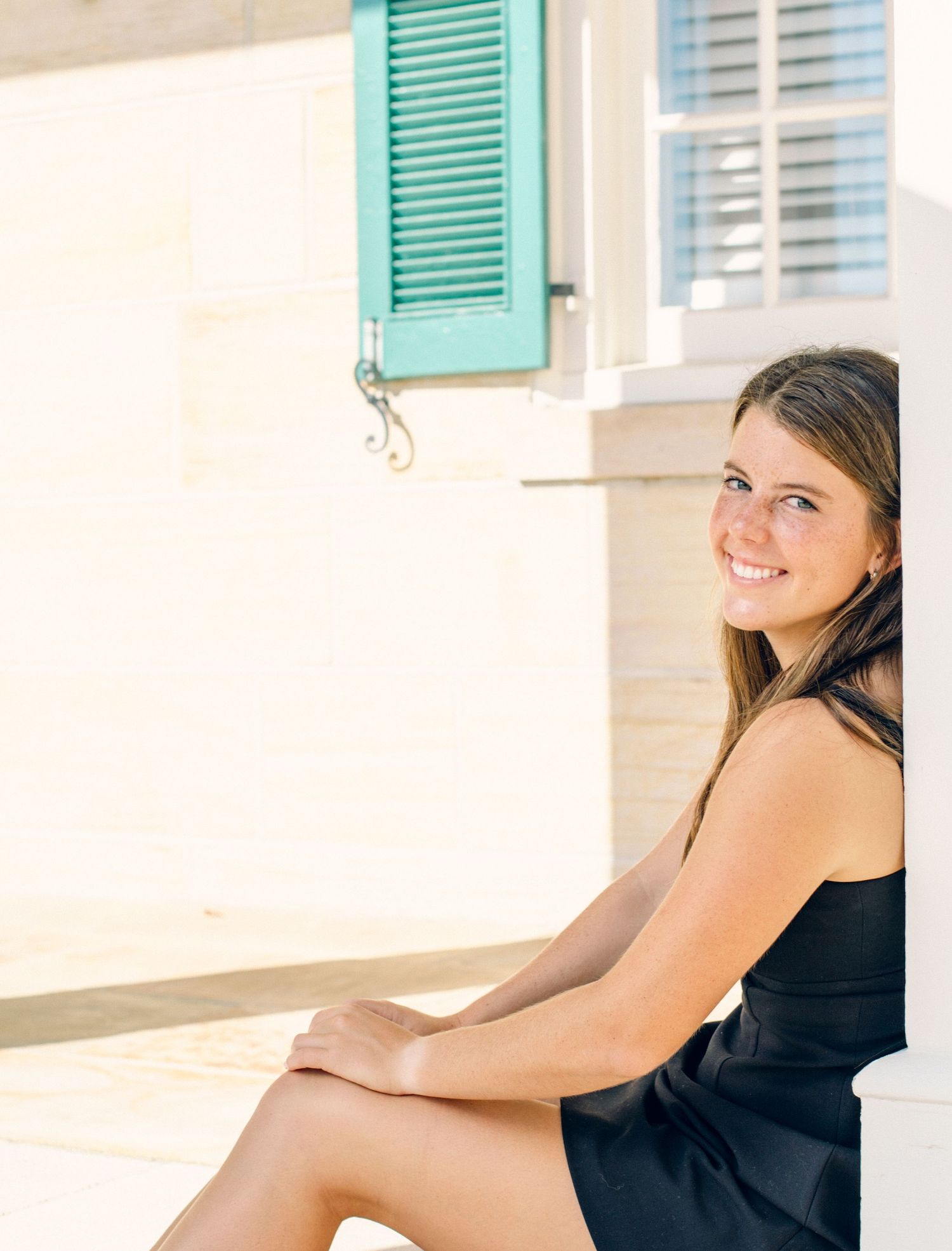 Girl in black dress sitting with bright blue shutter in background.