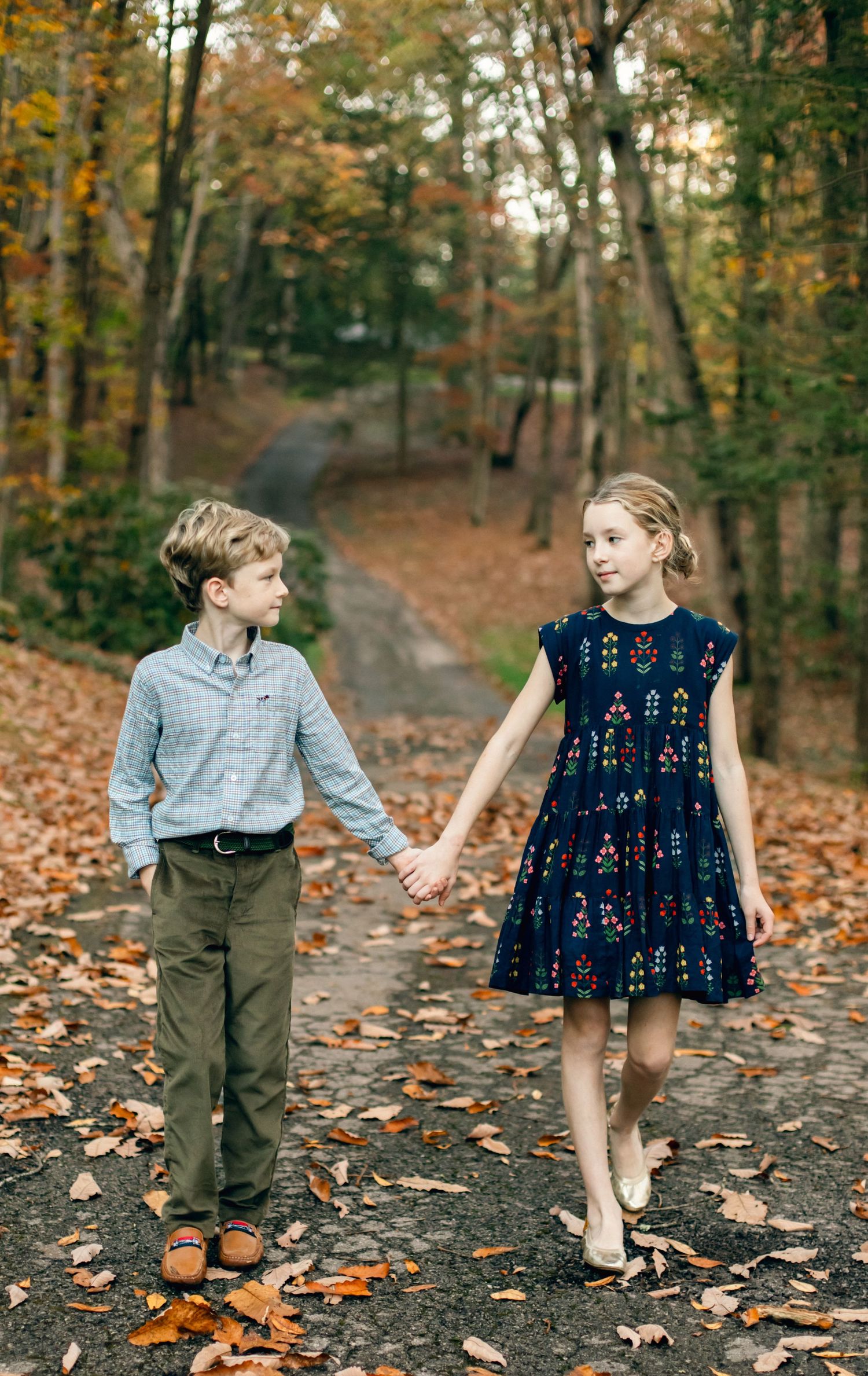 Two children holding hands on a leaf-covered autumn path, boy in blue shirt and girl in navy dress.