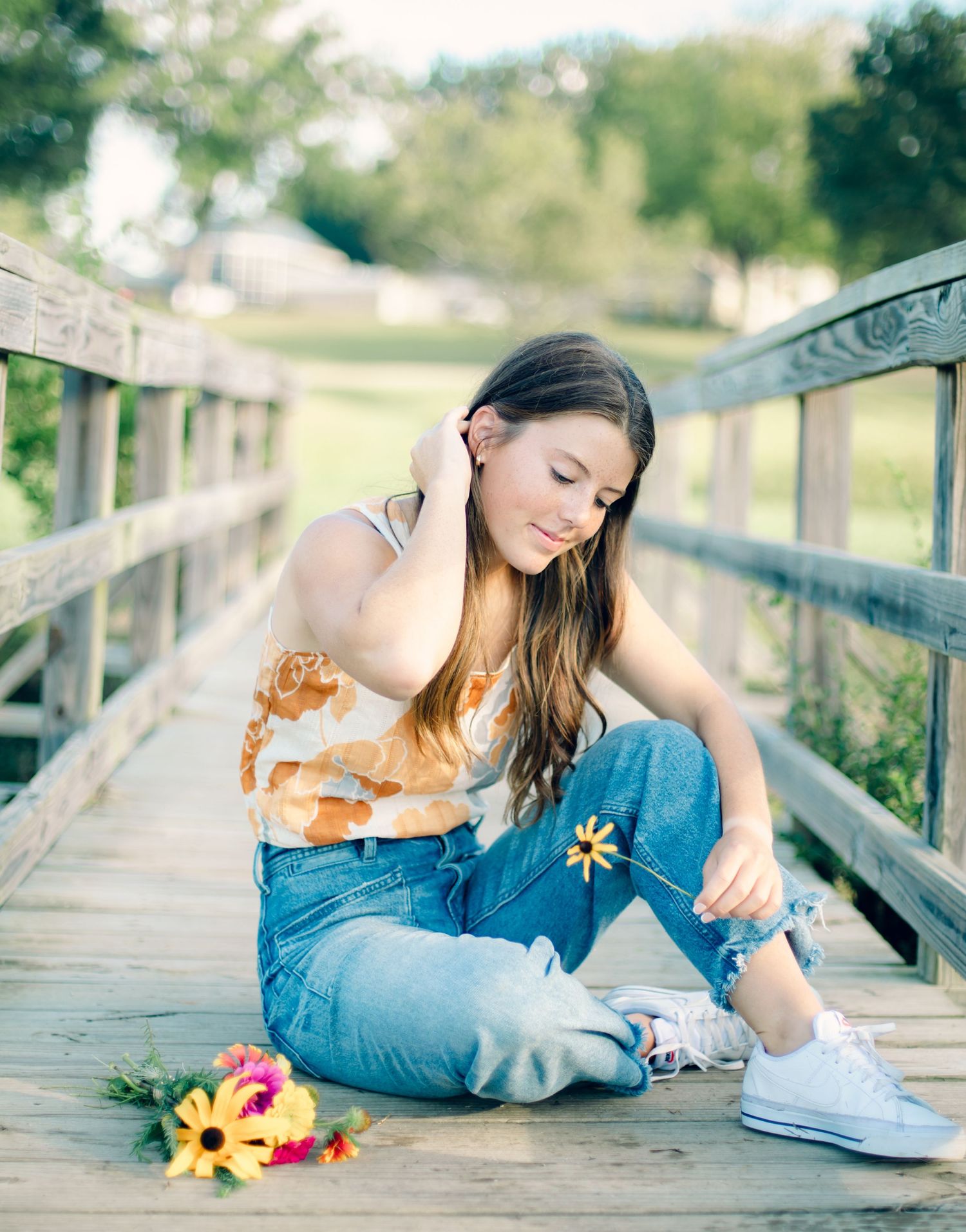 Young woman sitting on wooden bridge holding yellow daisy with colorful bouquet nearby, wearing jeans and floral top.