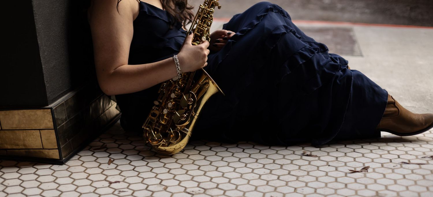 Woman in navy dress sitting on black and white hexagon tiled floor holding a gold saxophone against a brick wall.
