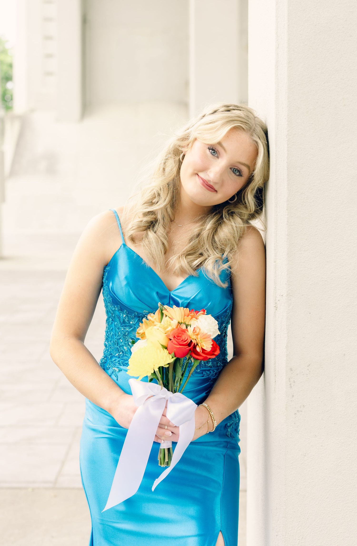 Blonde woman in blue satin gown holding colorful bouquet of roses, leaning against white pillar.