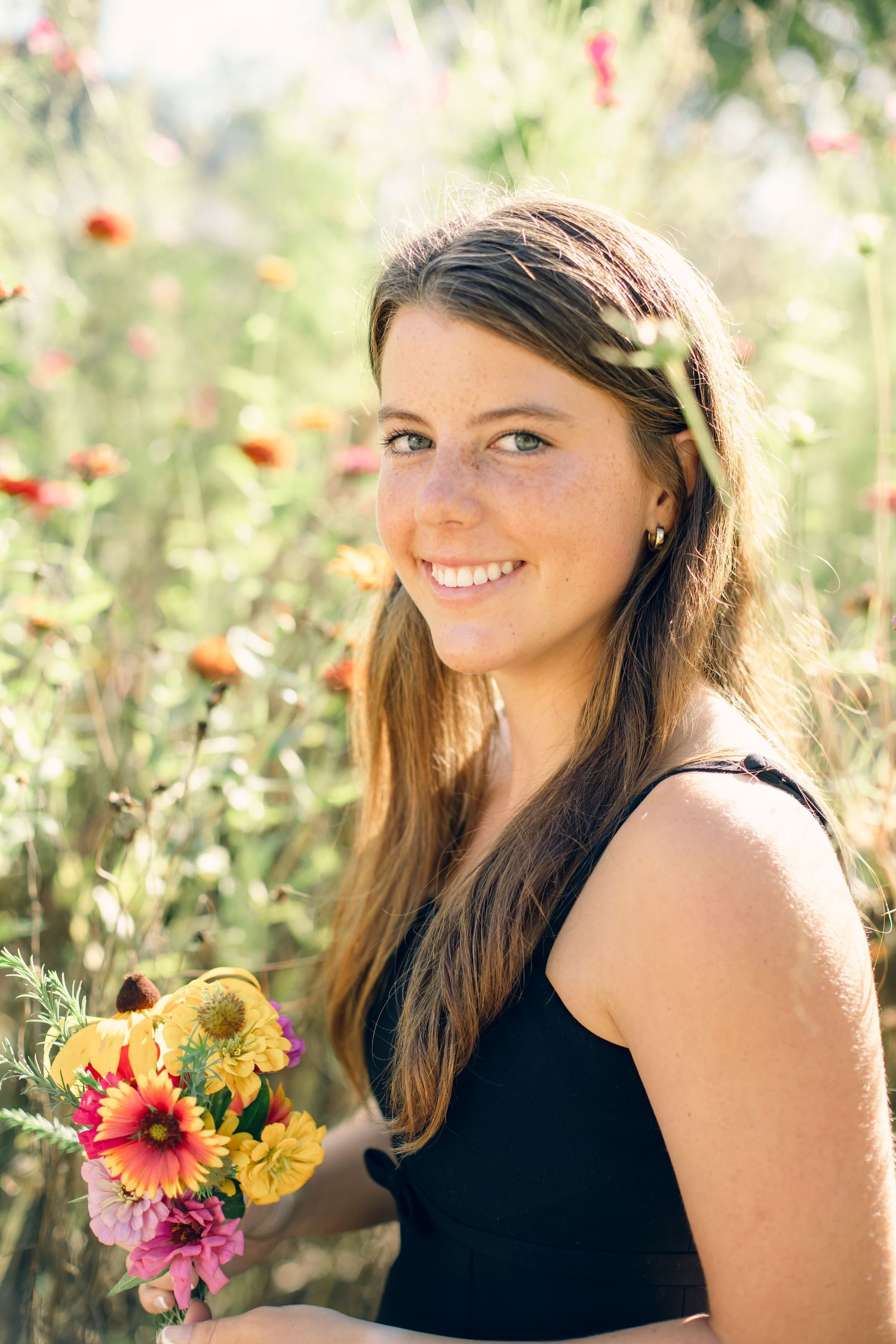Smiling young woman with freckles holding colorful wildflowers in a sunny outdoor field.