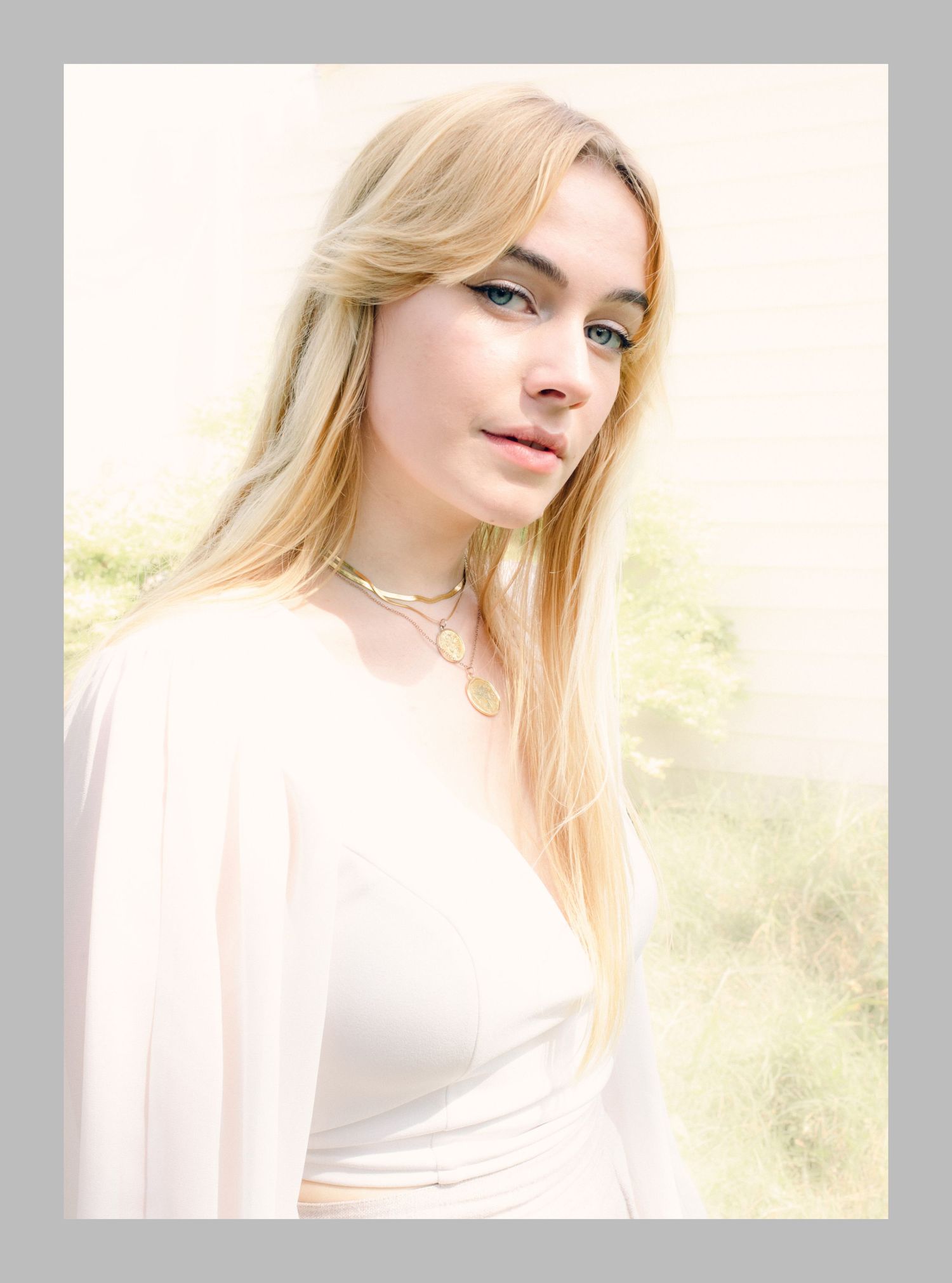 Young blonde woman in a white dress poses outdoors in a soft, dreamy light with blurred green trees behind her.