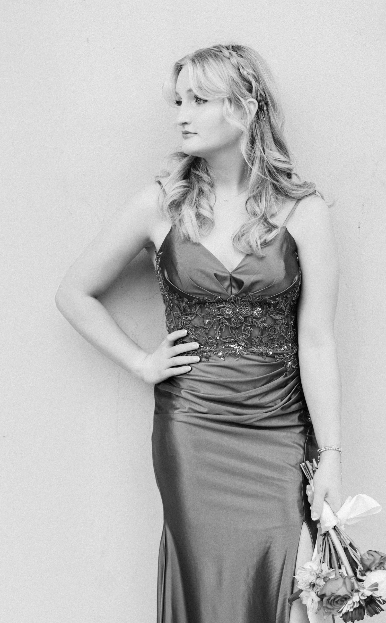 Black and white photo of Senior EK wearing a  satin gown holding a bouquet, posing gracefully against a wall.