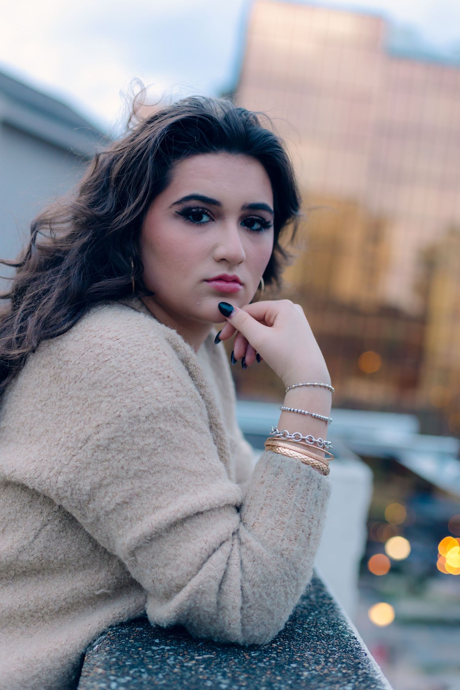 Chattanooga Senior girl with dark wavy hair and bold makeup wearing a beige sweater and gold bracelets posing outdoors at dusk.