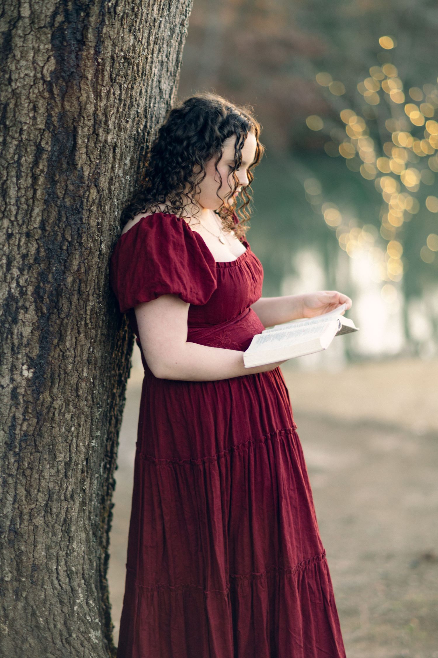 Young woman in red dress reads book leaning against tree in soft bokeh outdoor setting.