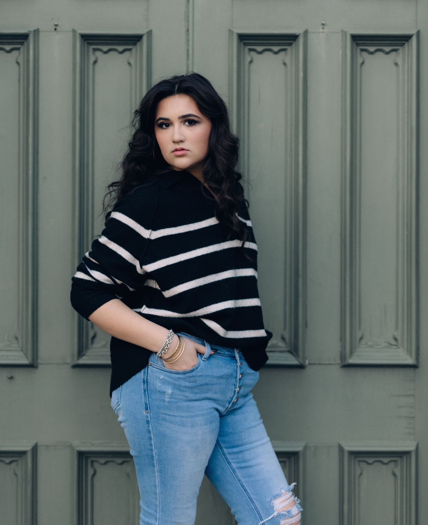Young woman in striped black sweater and ripped jeans posing against a sage green paneled door.